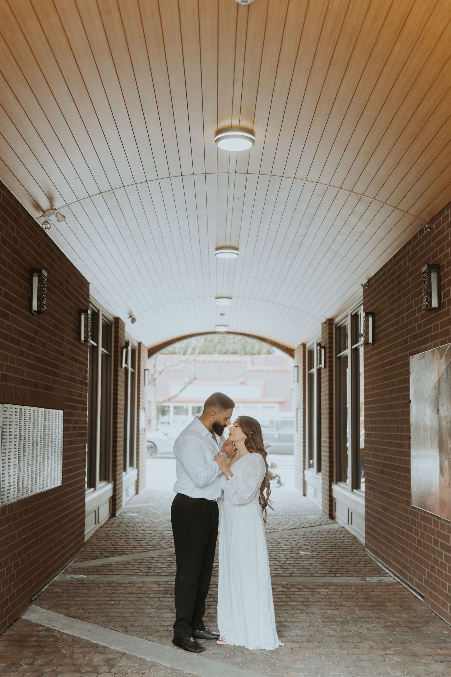 Two people in formal attire share a romantic moment in a covered brick walkway with white ceiling.