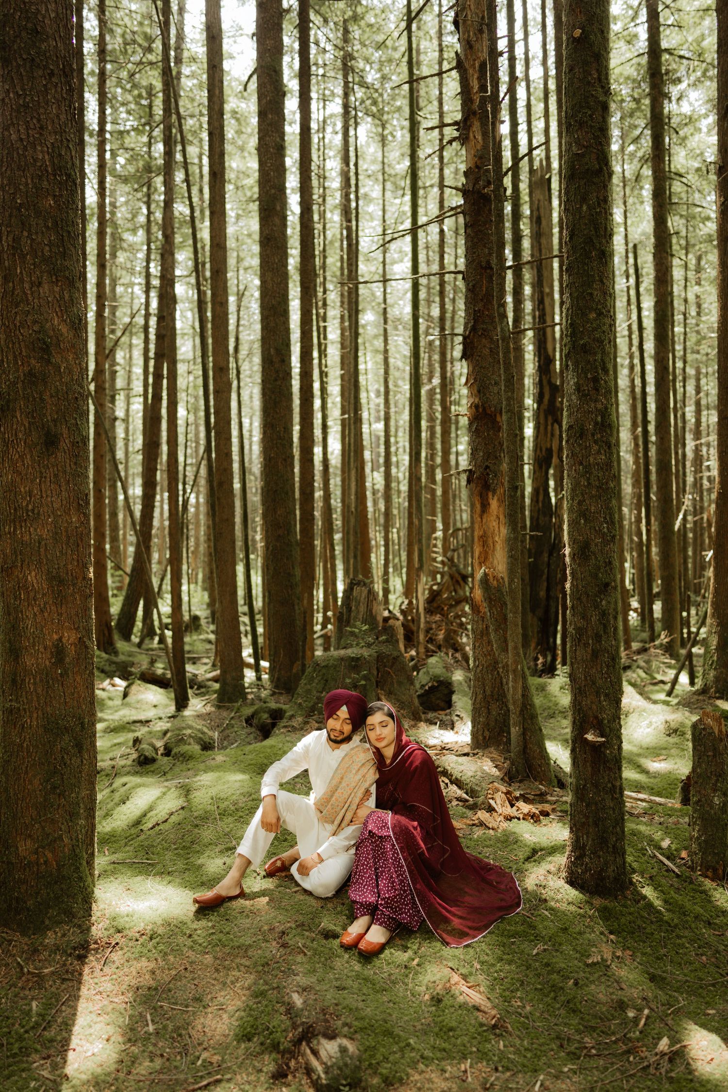 Sikh couple sitting together on moss-covered forest floor surrounded by tall pine trees.