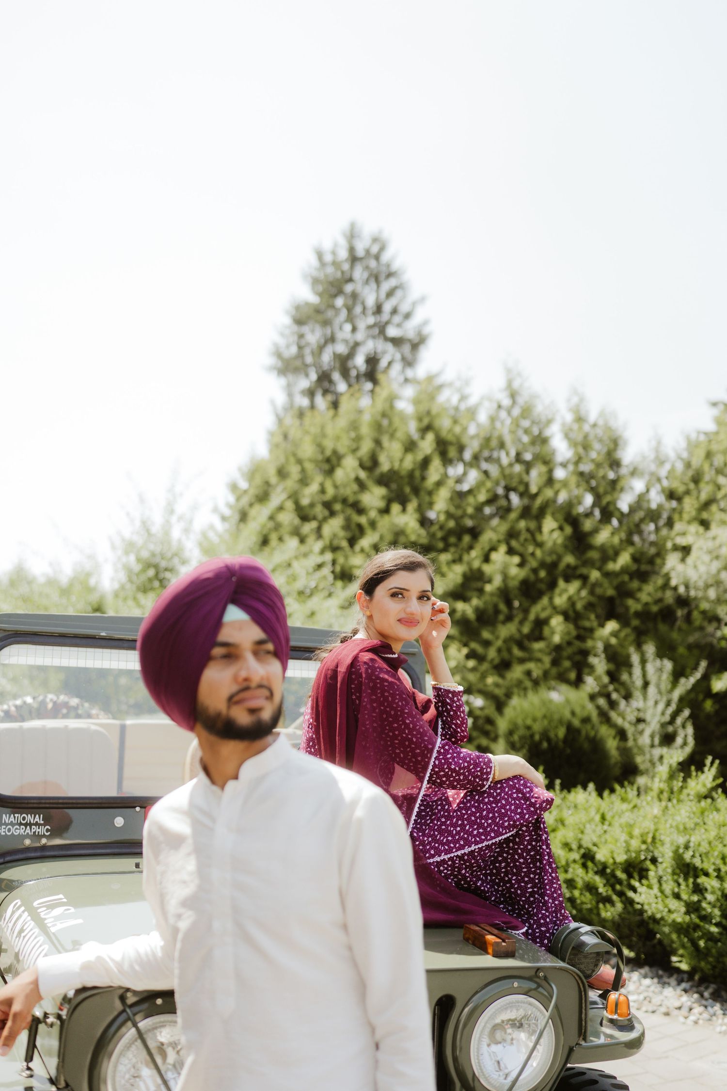 Sikh man in white kurta and purple turban stands by jeep as woman in maroon salwar sits on hood.