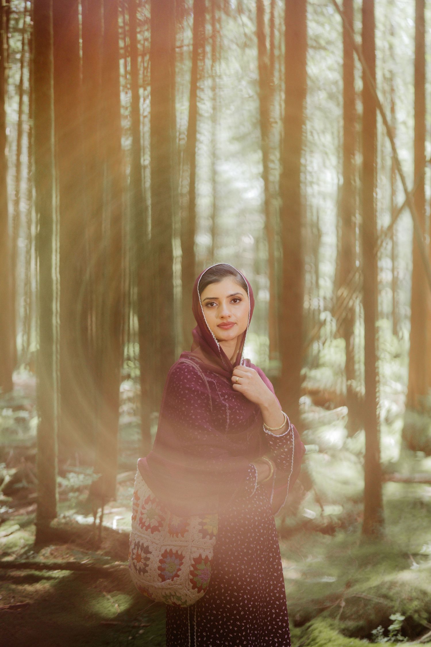 South Asian woman in burgundy traditional dress with head covering poses in a dreamy sunlit forest.