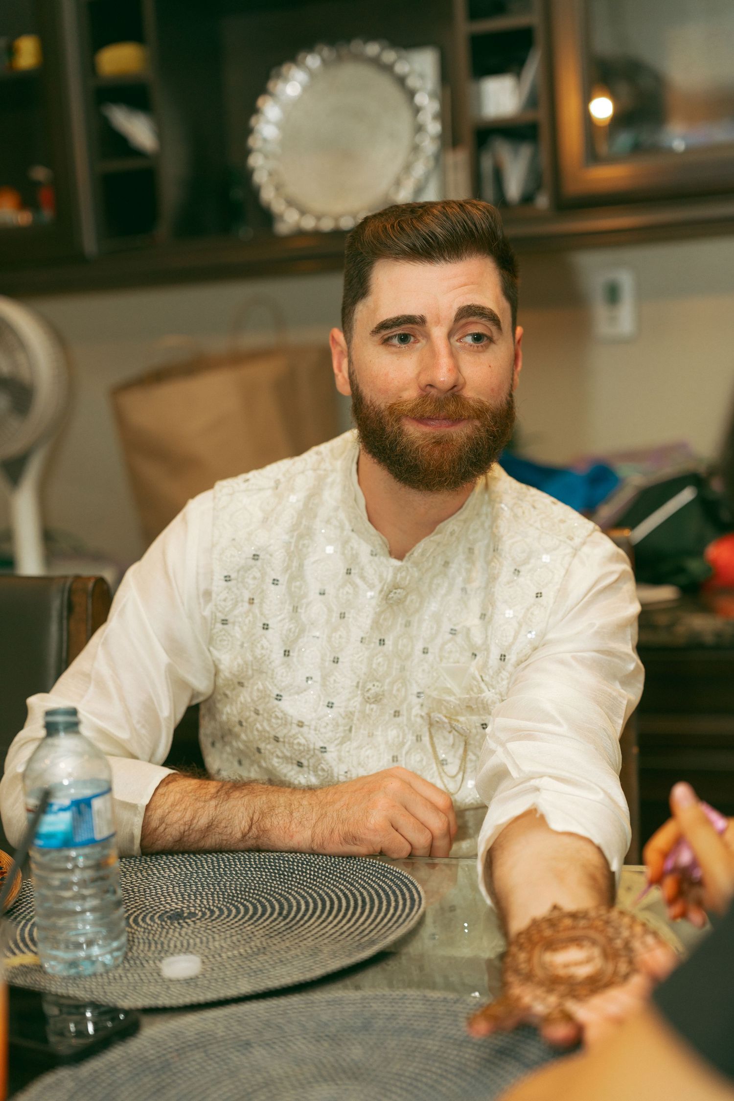 Bearded man in white embroidered kurta sits at glass table with water bottle, smiling warmly at a mehndi ceremony.