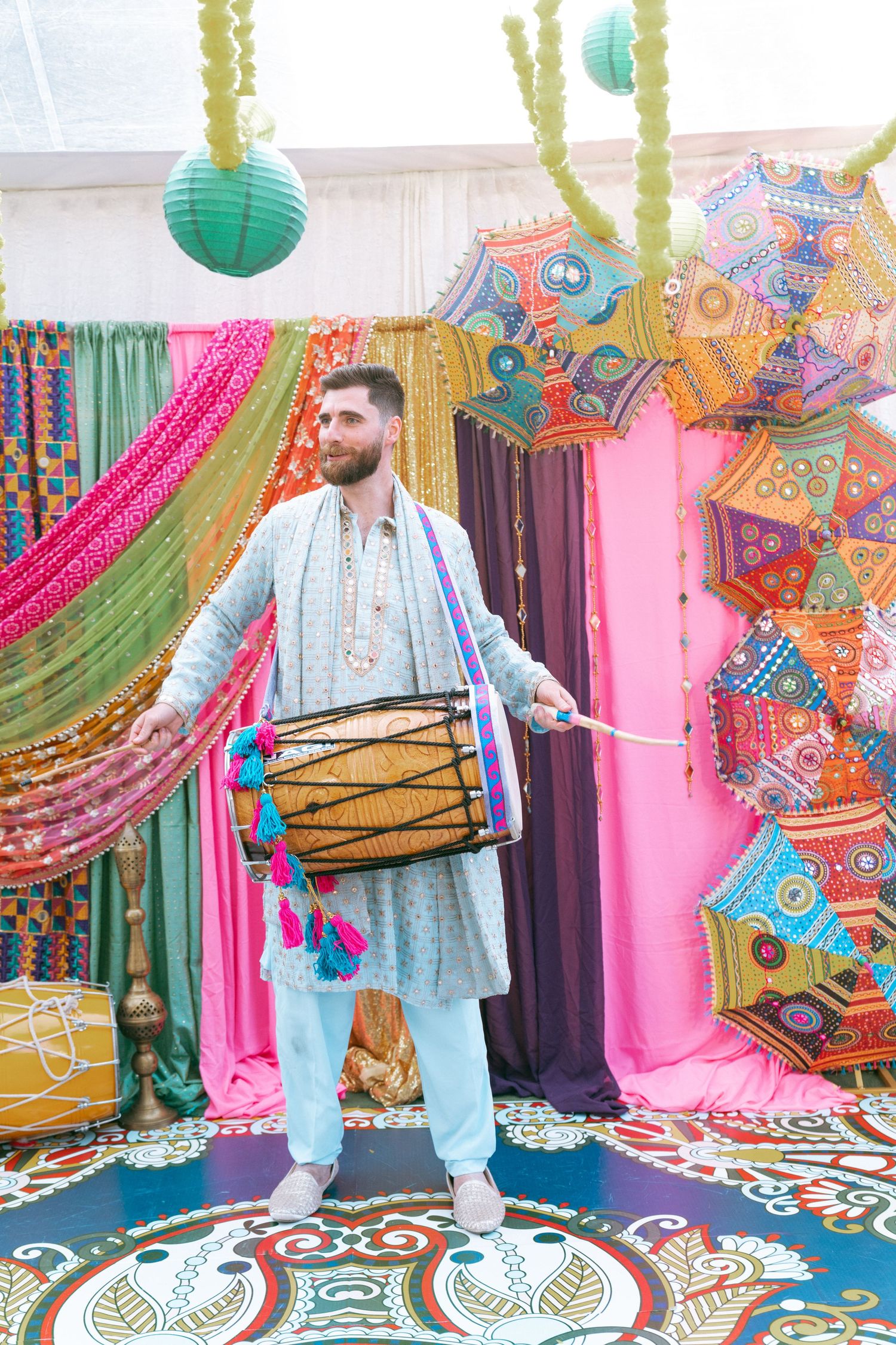 Man in traditional Indian kurta playing dhol drum at colorful Mehndi celebration with decorative umbrellas.