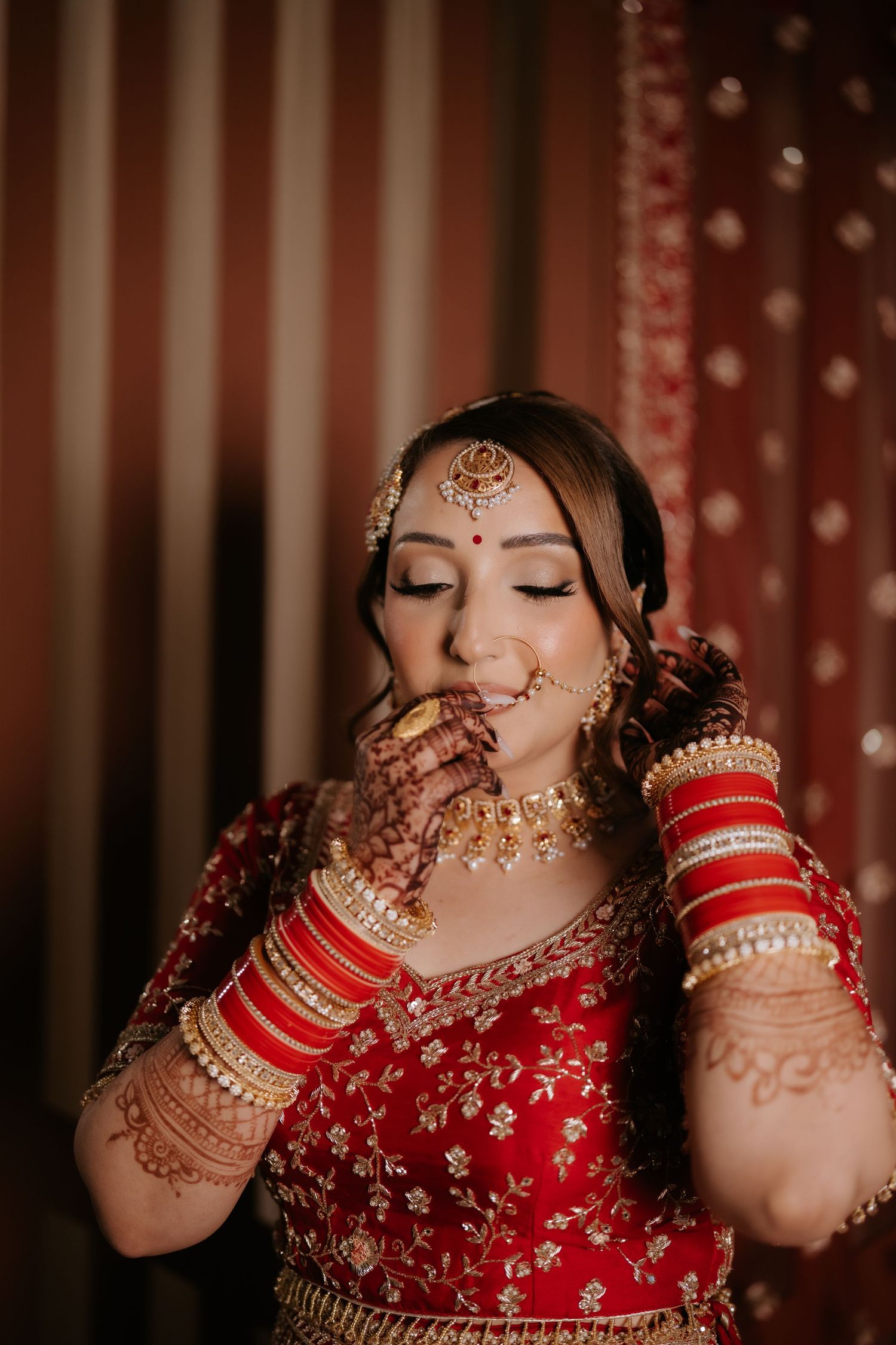 Indian bride in red lehenga with gold jewelry, mehndi hands, and red bangles posing elegantly at wedding ceremony.