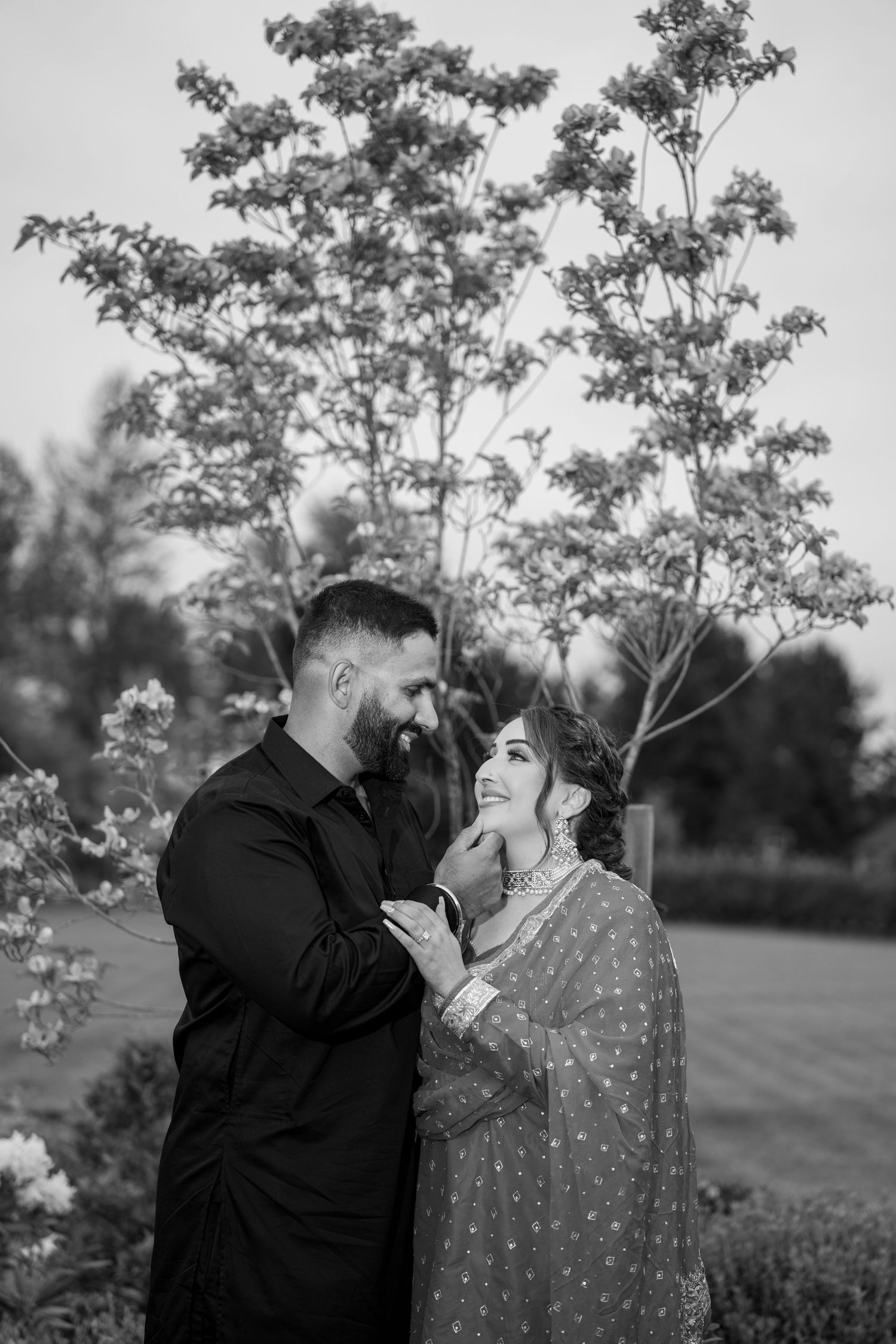 Black and white photo of a South Asian couple in traditional attire sharing a romantic moment outdoors near blooming trees.