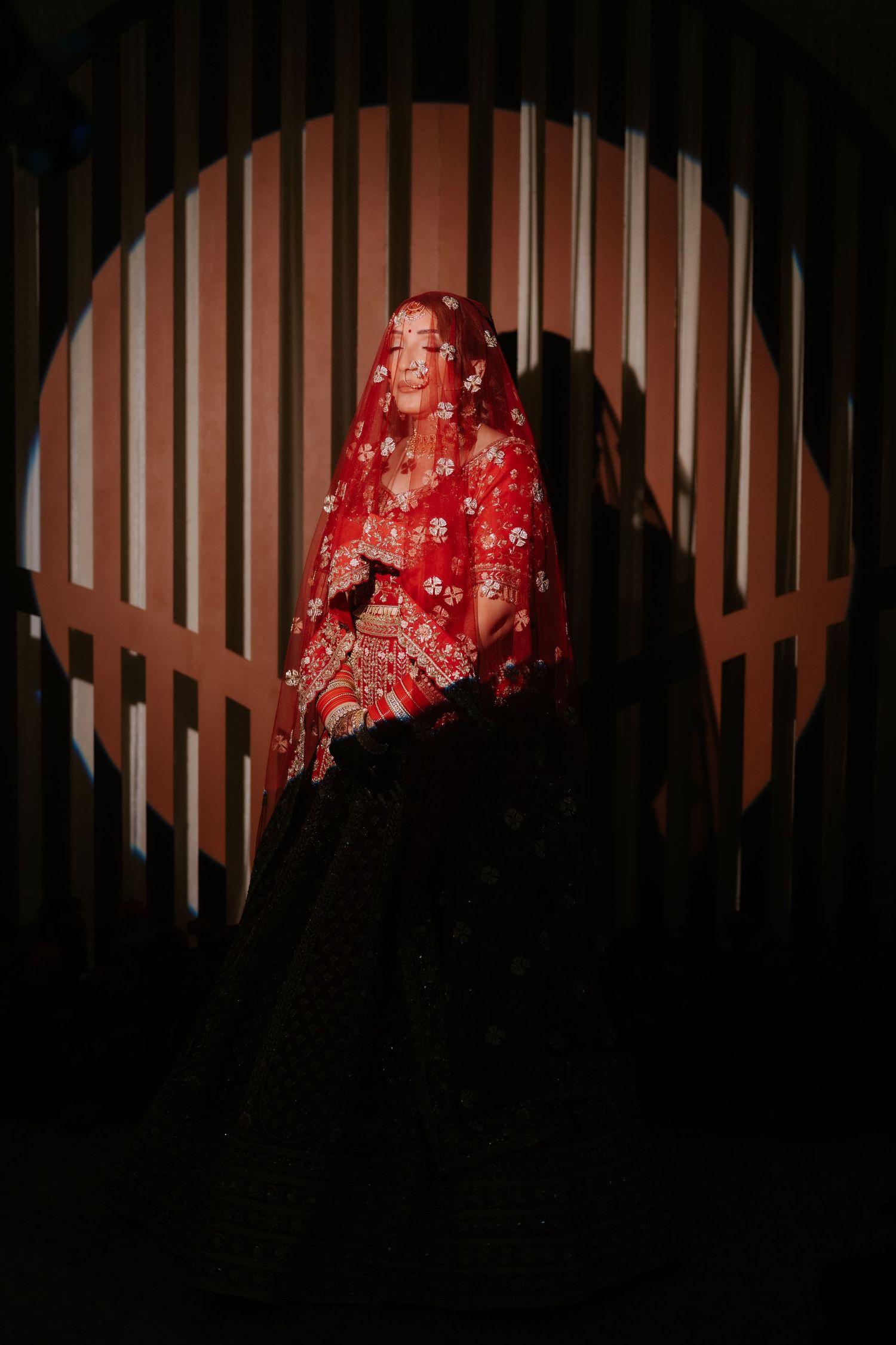 Bride in red embroidered lehenga and dupatta veil, posed in dramatic circular arch with striped shadow backdrop.