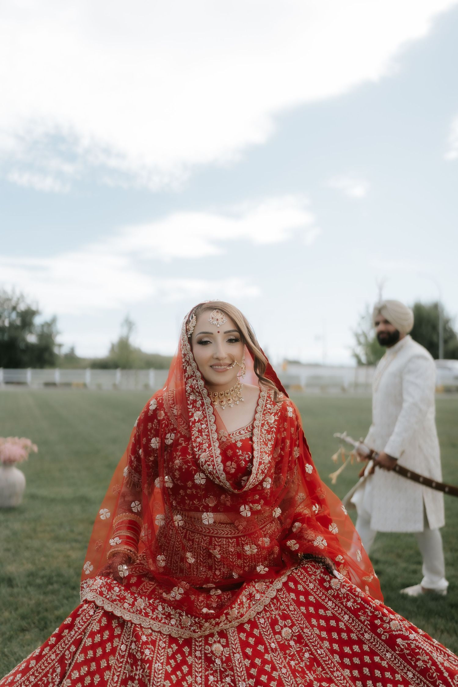Sikh bride in red lehenga with gold embroidery and dupatta smiles outdoors, groom in white sherwani stands behind.