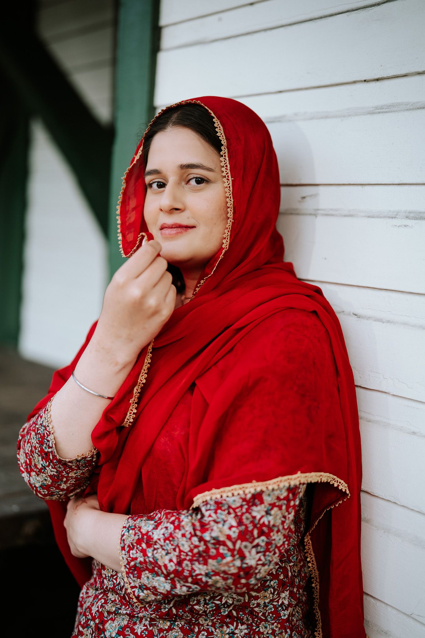 Close-up portrait of a smiling South Asian woman in red dupatta with gold trim against a white wooden wall.