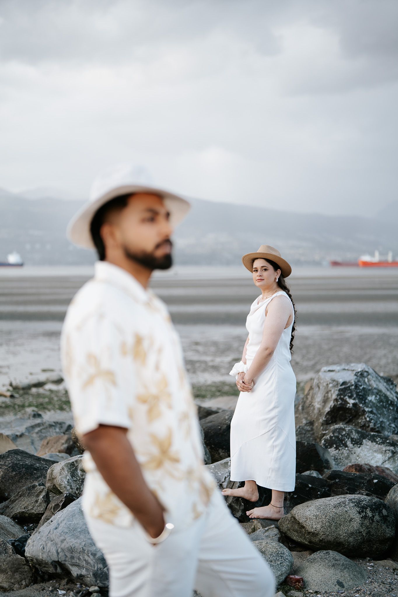 Couple in white outfits and hats pose on rocky beach with ocean and mountains in background.