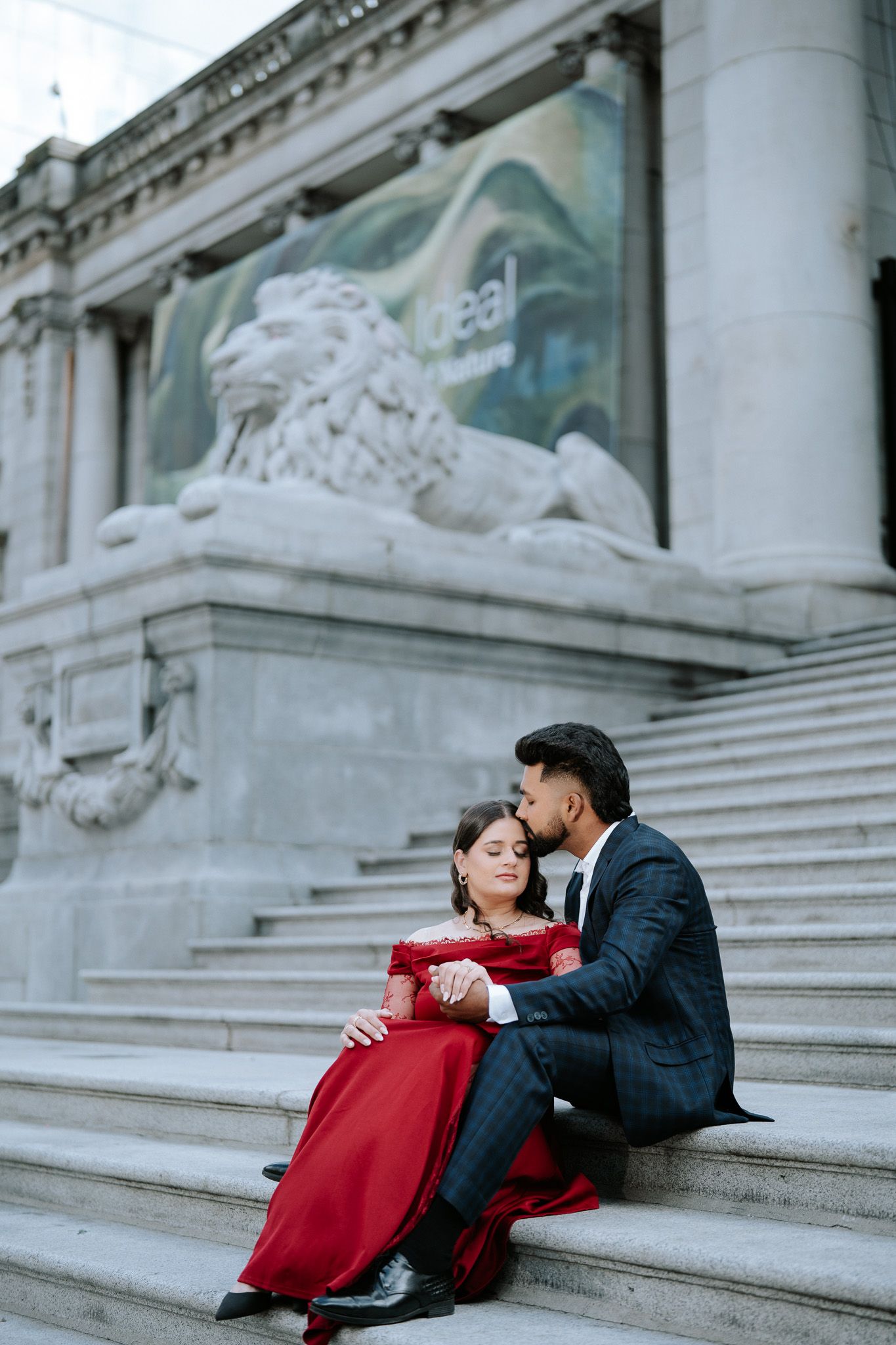 Couple in elegant red dress and suit sitting on steps of neoclassical building with lion statue.