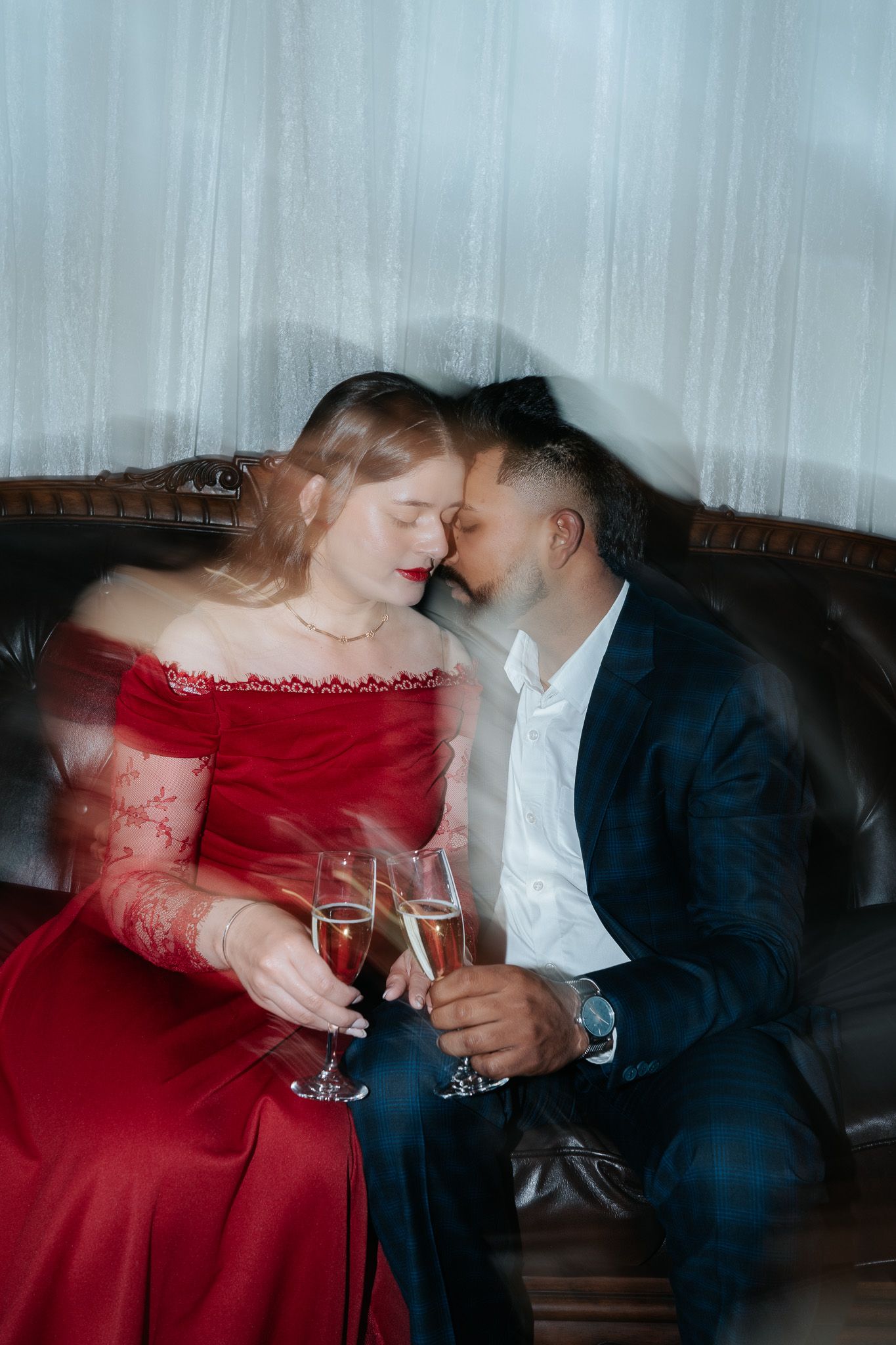 Romantic couple in elegant attire sharing champagne on a leather sofa, woman in red dress, man in navy suit.