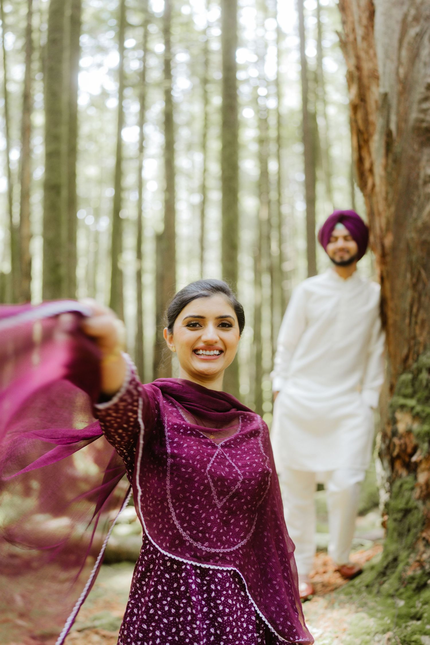 A smiling woman in purple salwar kameez twirls her dupatta in a forest with a man in background.