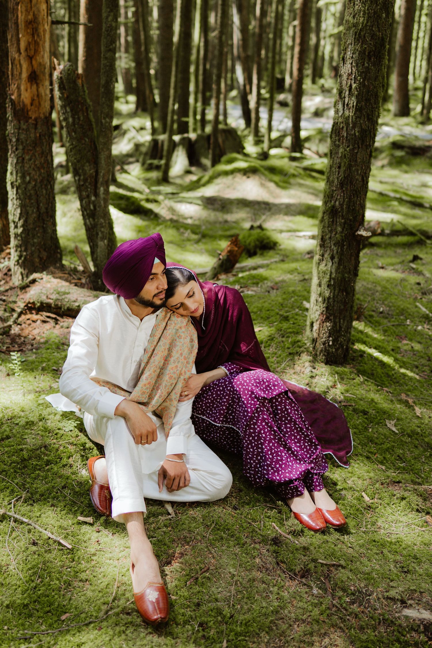 Sikh couple in purple and white traditional outfits sitting on moss in a lush forest.
