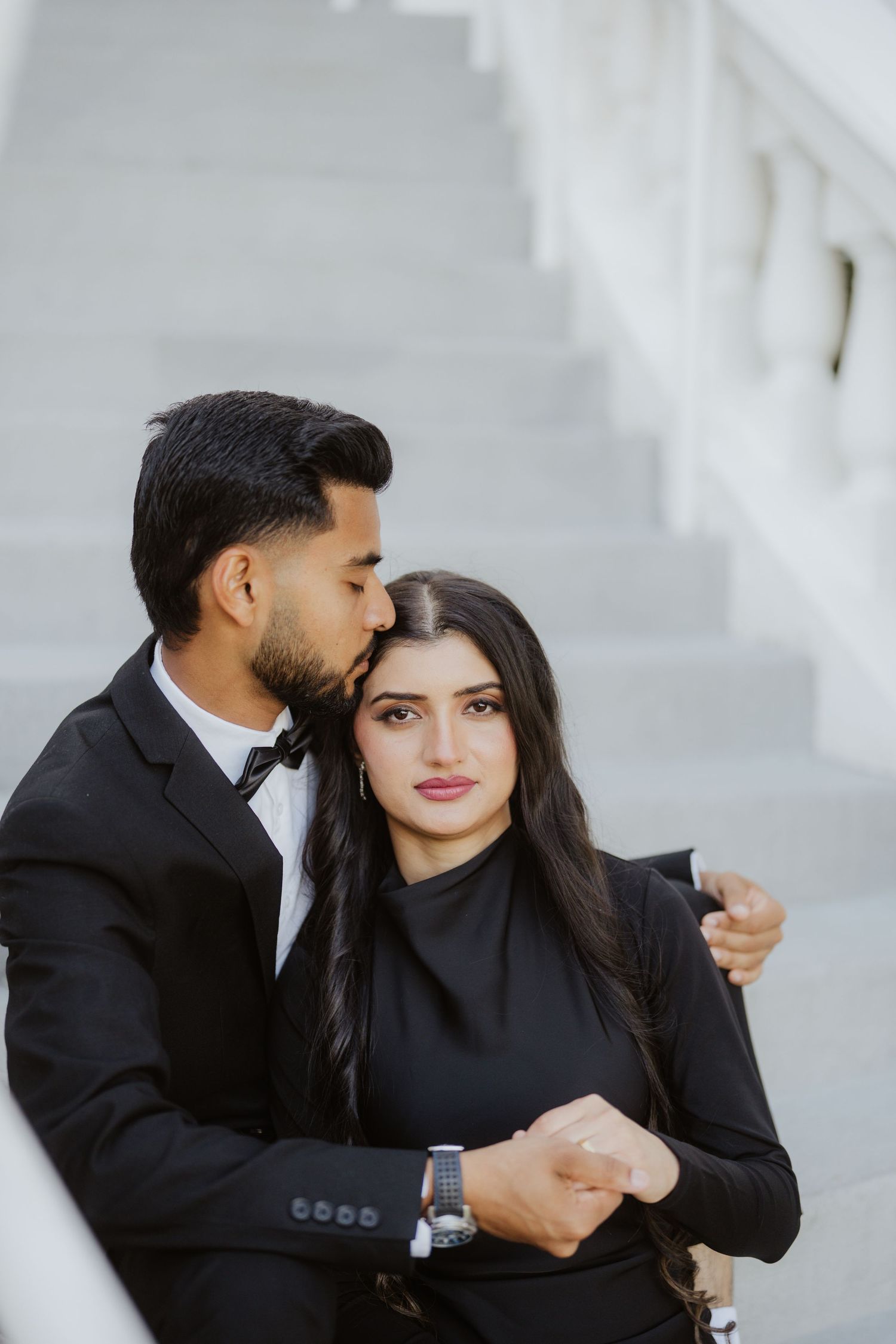 South Asian man in black suit kissing woman in black dress on the cheek while seated on white marble steps.