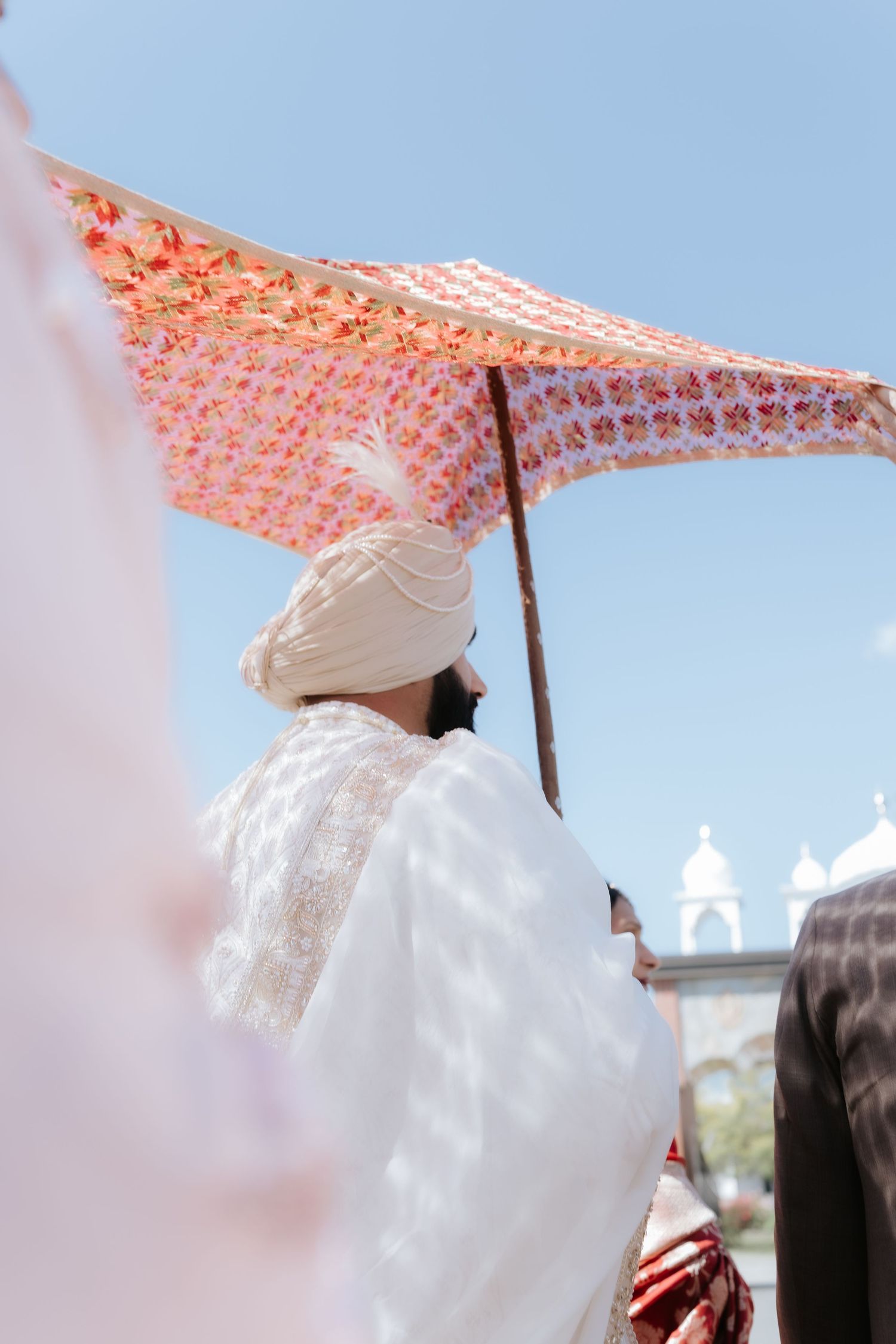 Sikh groom in white sherwani and turban walks under decorative floral canopy during outdoor wedding ceremony.