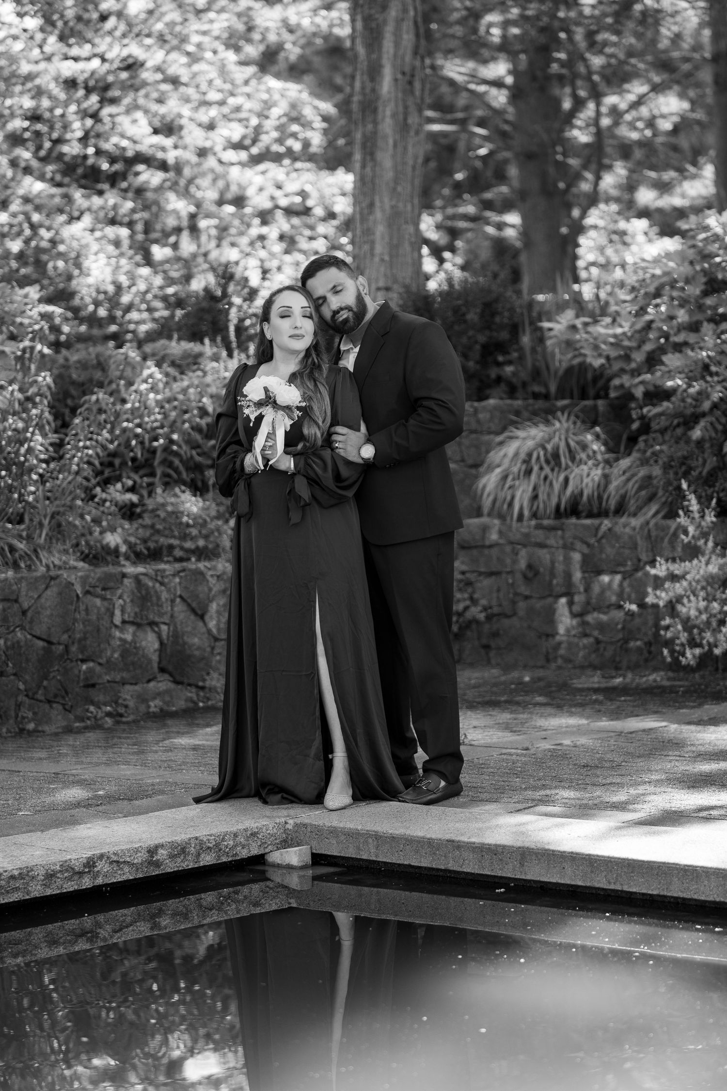 Black and white photo of a couple in formal attire posing by a reflective pool in a lush garden setting.
