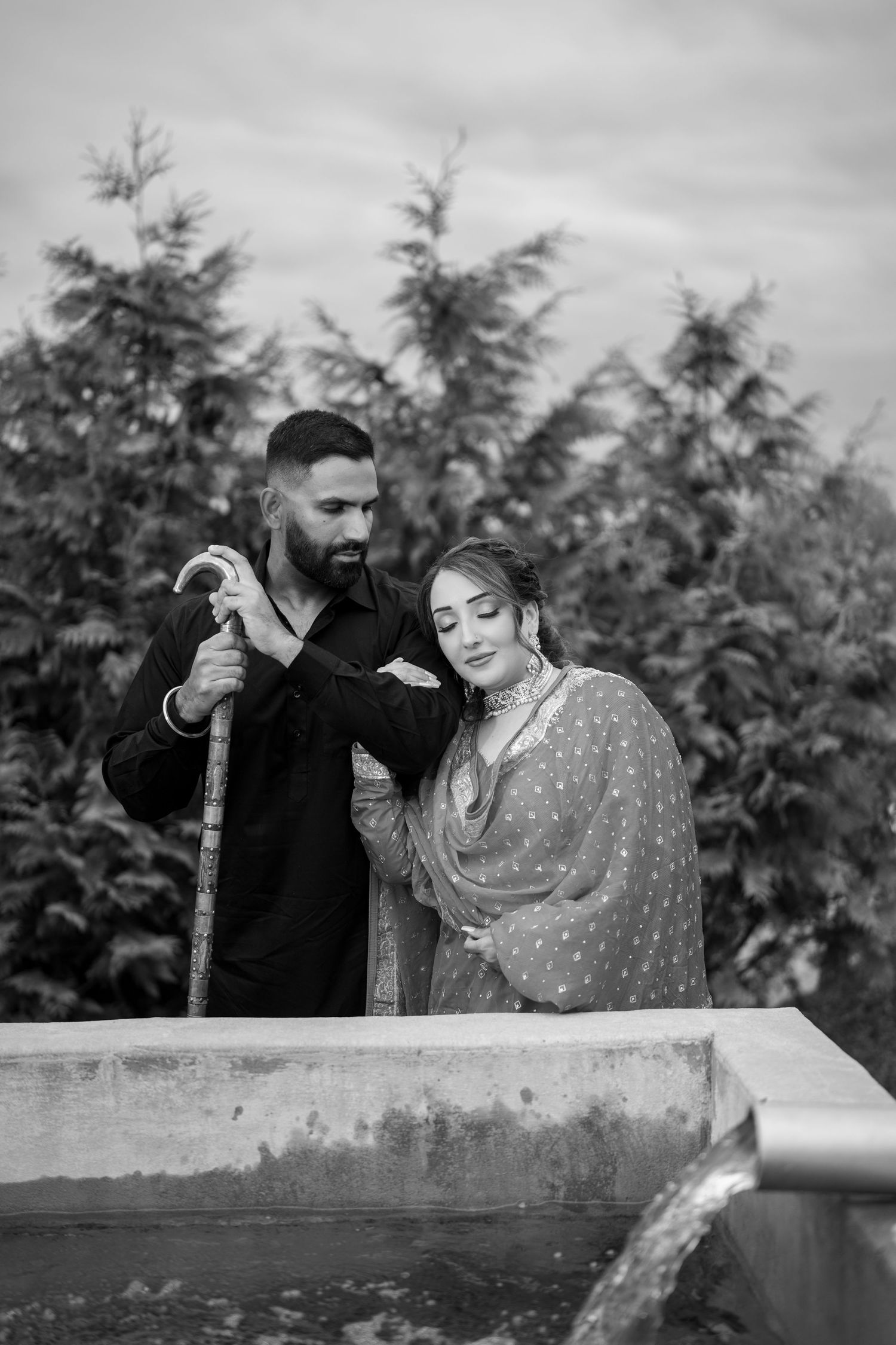 Black and white photo of a bearded South Asian man in black and a woman in traditional salwar kameez posing near a well outdoors.