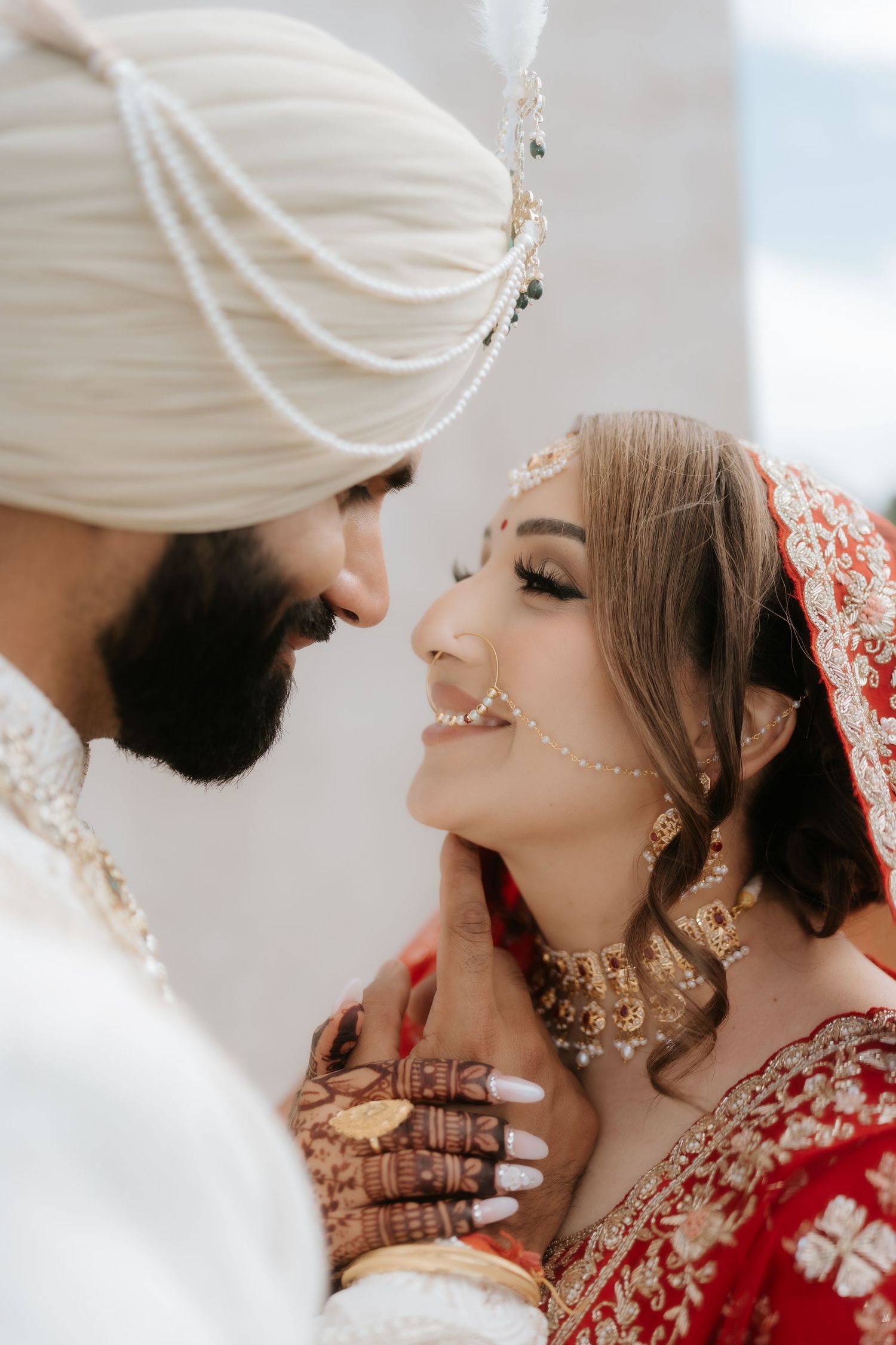 A Sikh groom in white turban tenderly holds his bride's face, she wears red bridal lehenga with gold jewelry and mehndi.
