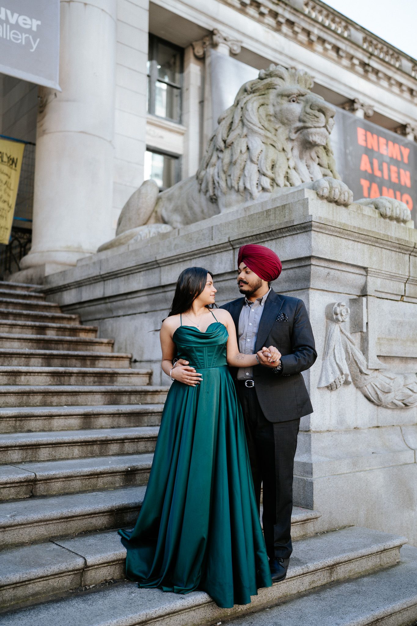 A couple in formal attire stands on stone steps by a lion statue, with the woman wearing an elegant emerald green gown.