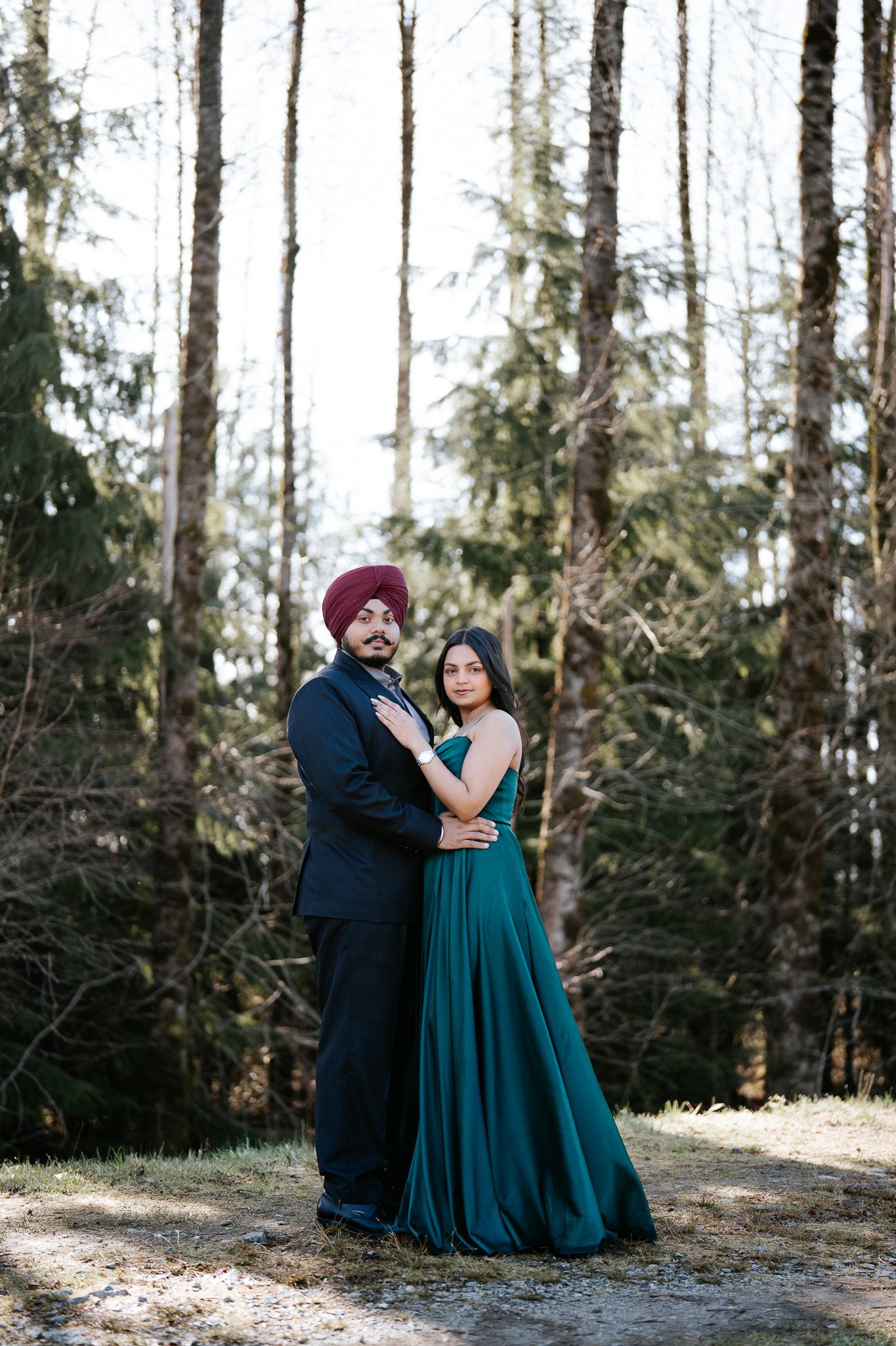 A couple shares romantic moments in a forest setting wearing formal attire against evergreen trees.
