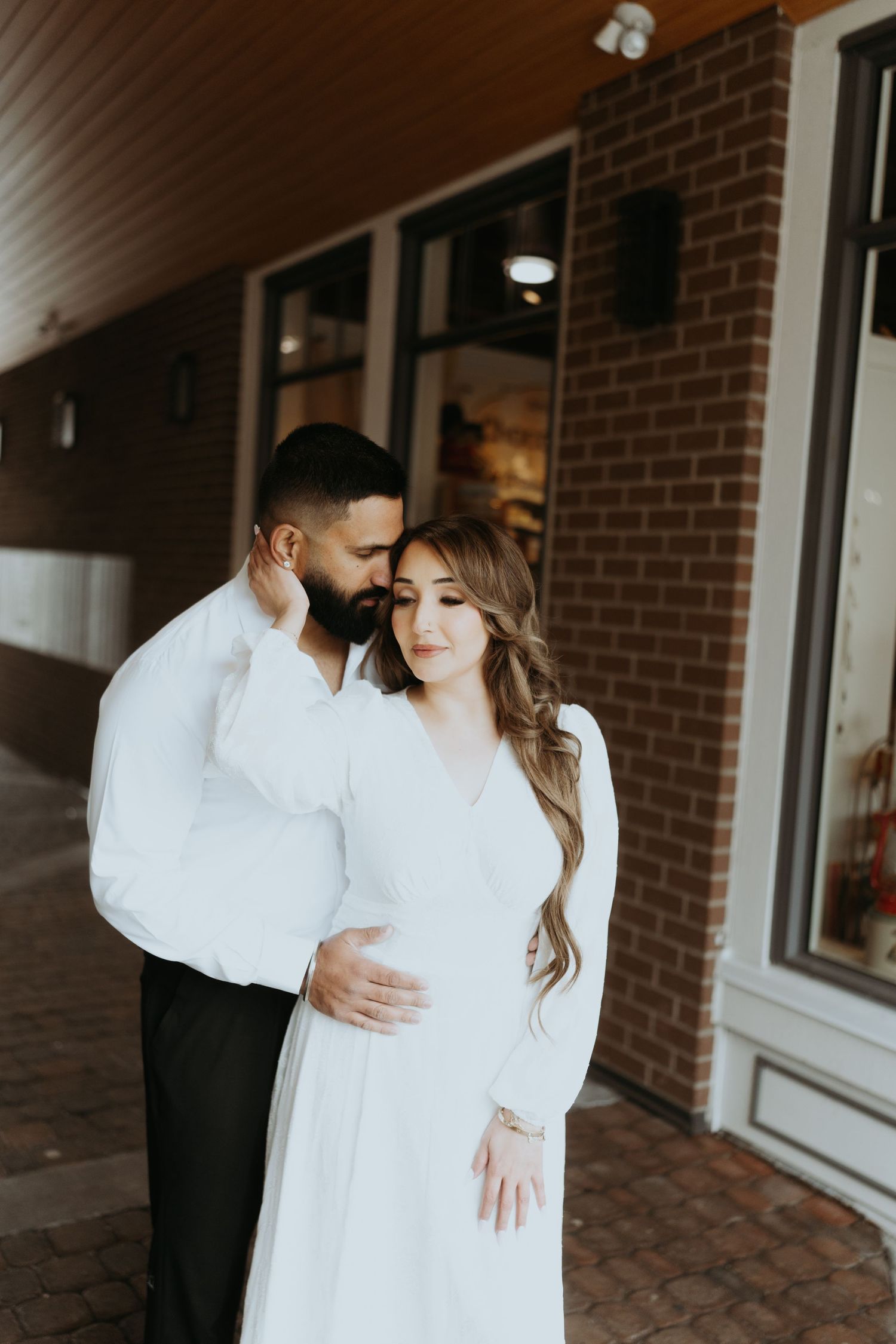 A couple in white clothing embraces in an intimate moment against a brick wall backdrop.