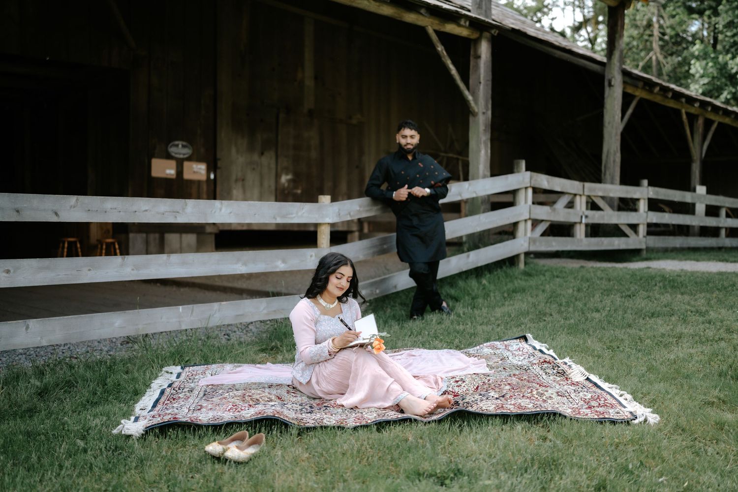 A couple enjoys a picnic setup with a rustic barn backdrop, sitting on a patterned blanket.