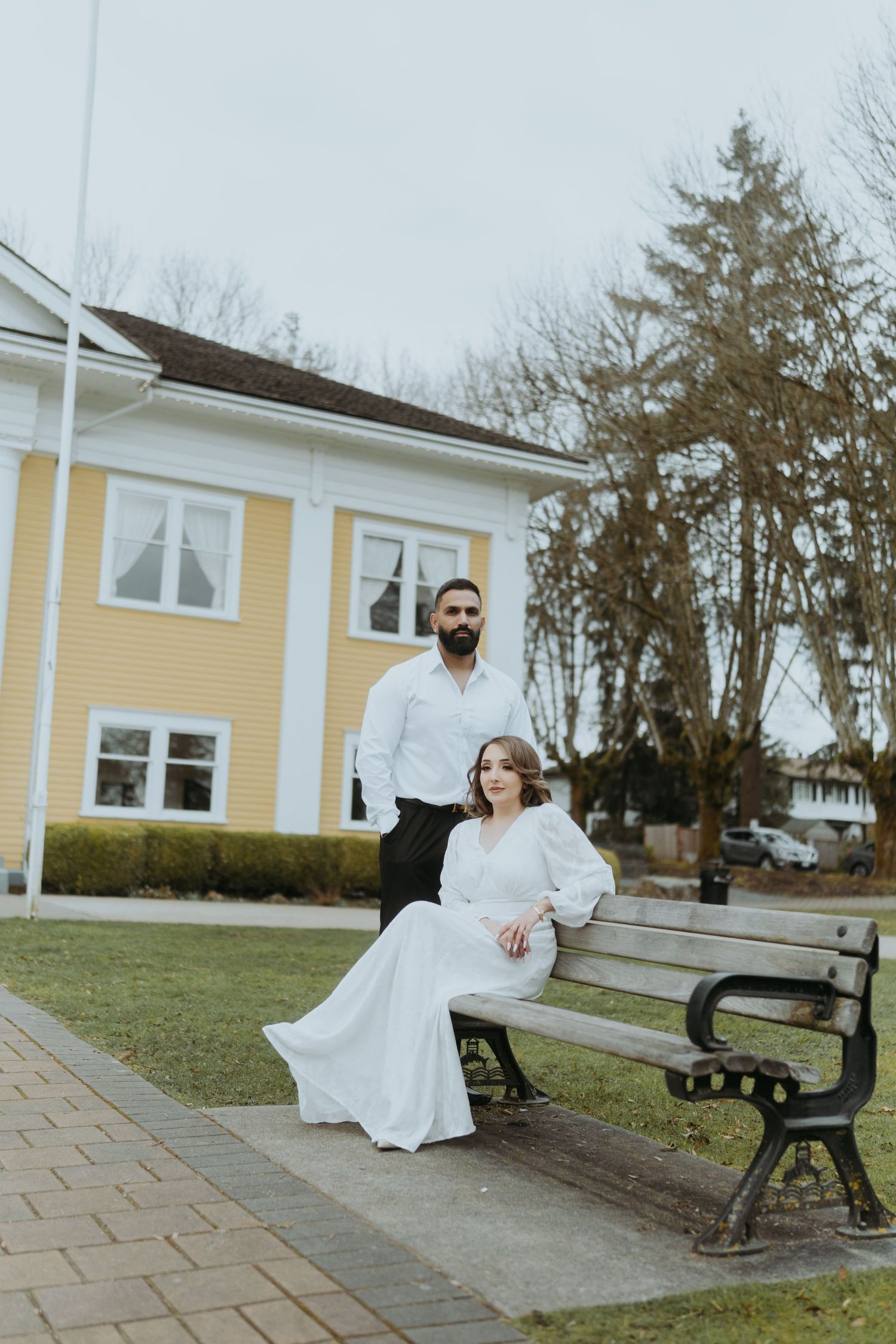 A couple shares a moment together on a bench outside a yellow house with white trim.
