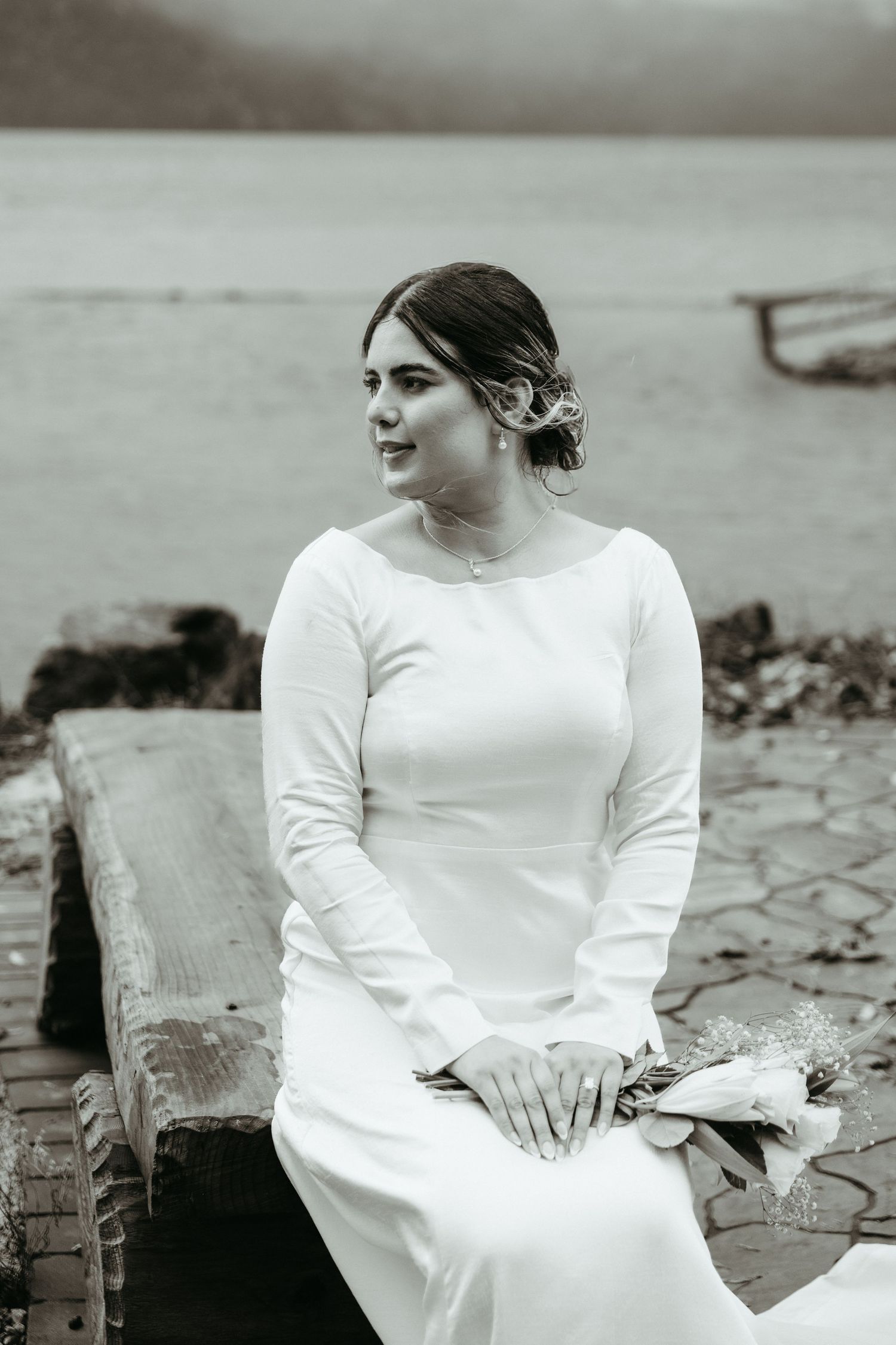 A person in a white long-sleeved dress sits thoughtfully by the water on a cloudy day in black and white.