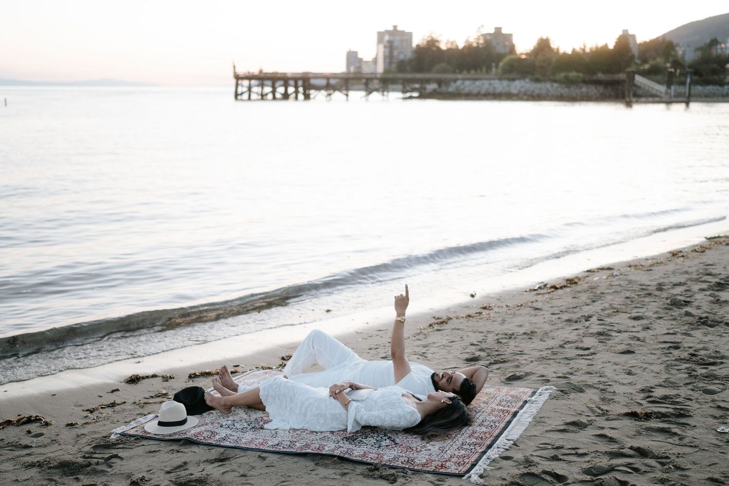 A person lies comfortably on a blanket at the beach during sunset with mountains visible in the background.