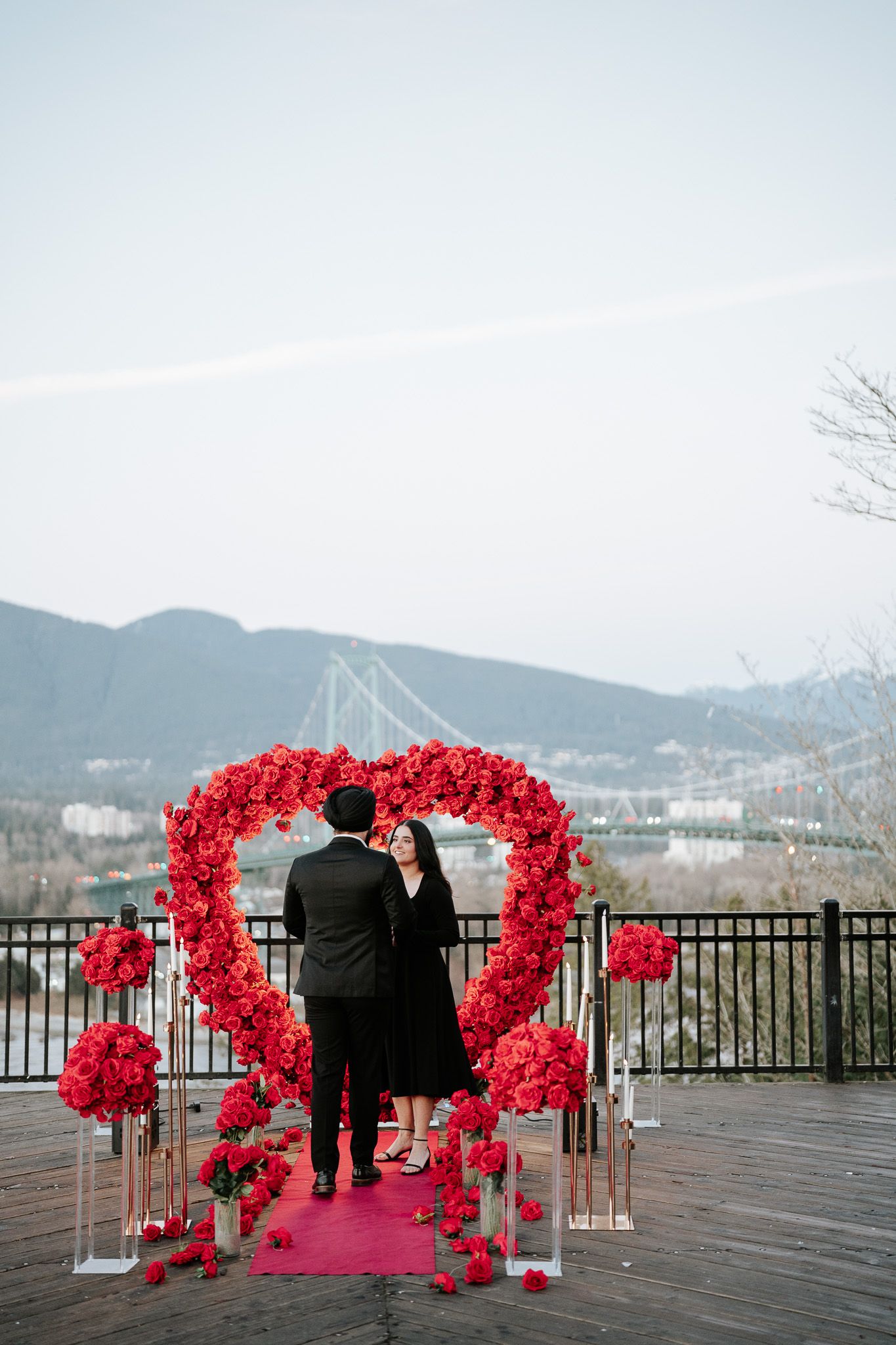 A romantic proposal setup on a waterfront deck with red floral arches and mountains in the background.