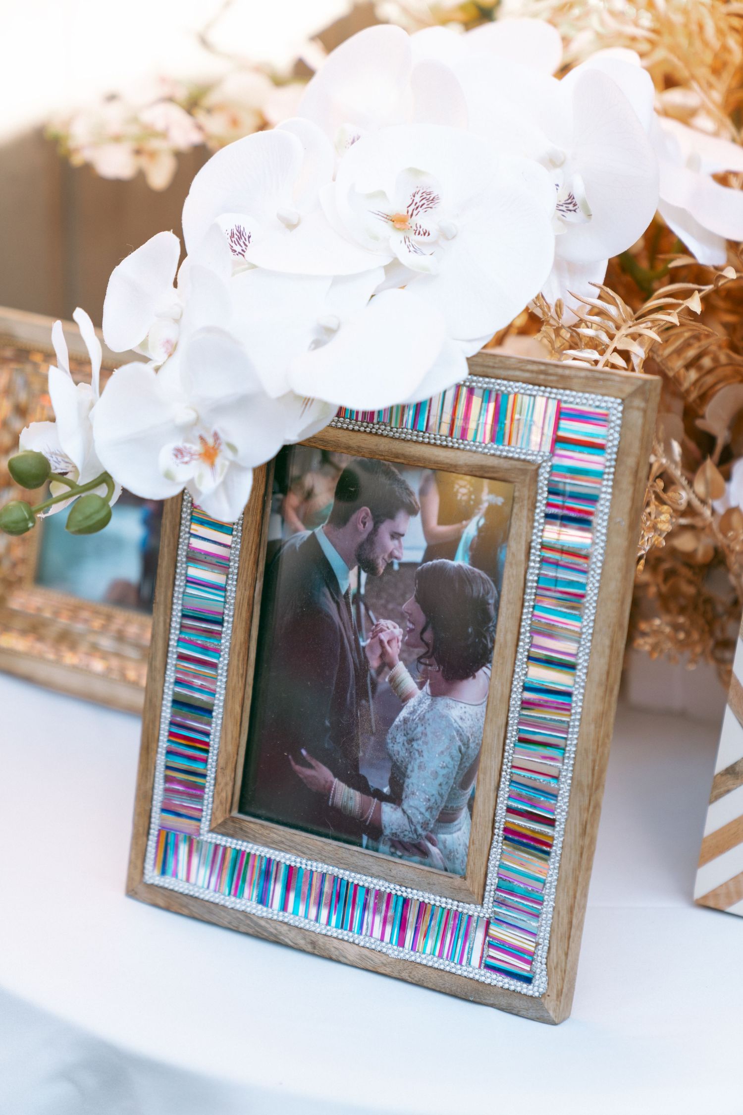 Colorful mosaic picture frame with white orchids displaying a wedding couple photo on a white table.