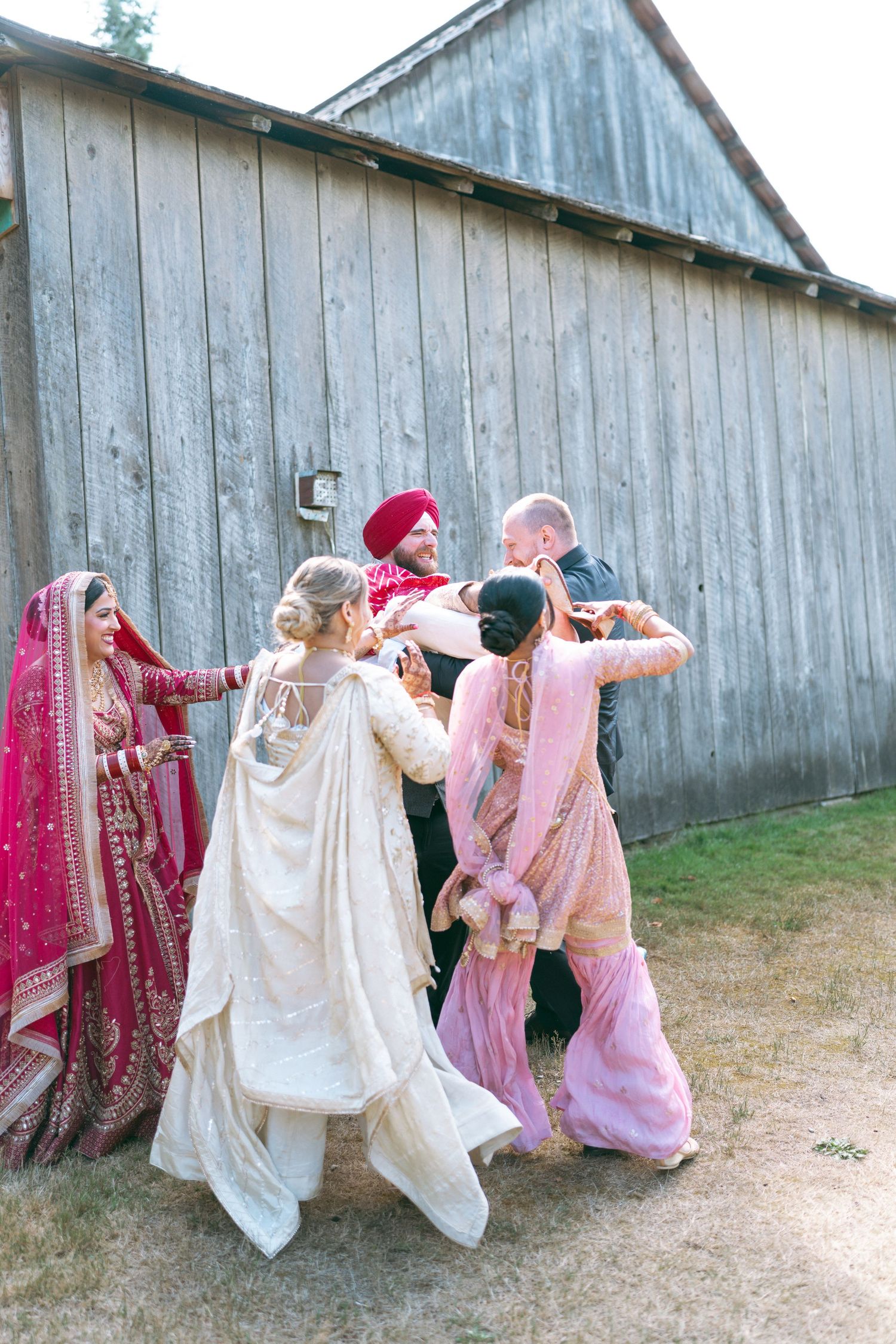 Wedding party in colorful Indian attire celebrates near a rustic barn, blending traditional and Western styles.