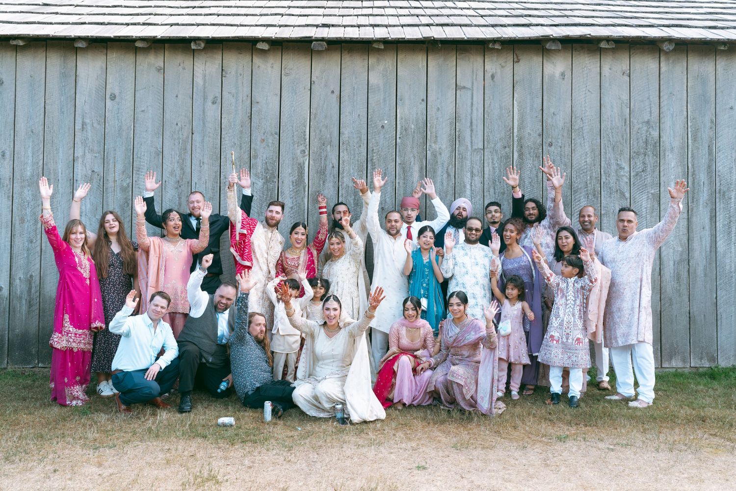 A joyful wedding group photo with guests in colorful traditional attire celebrating in front of a rustic barn.
