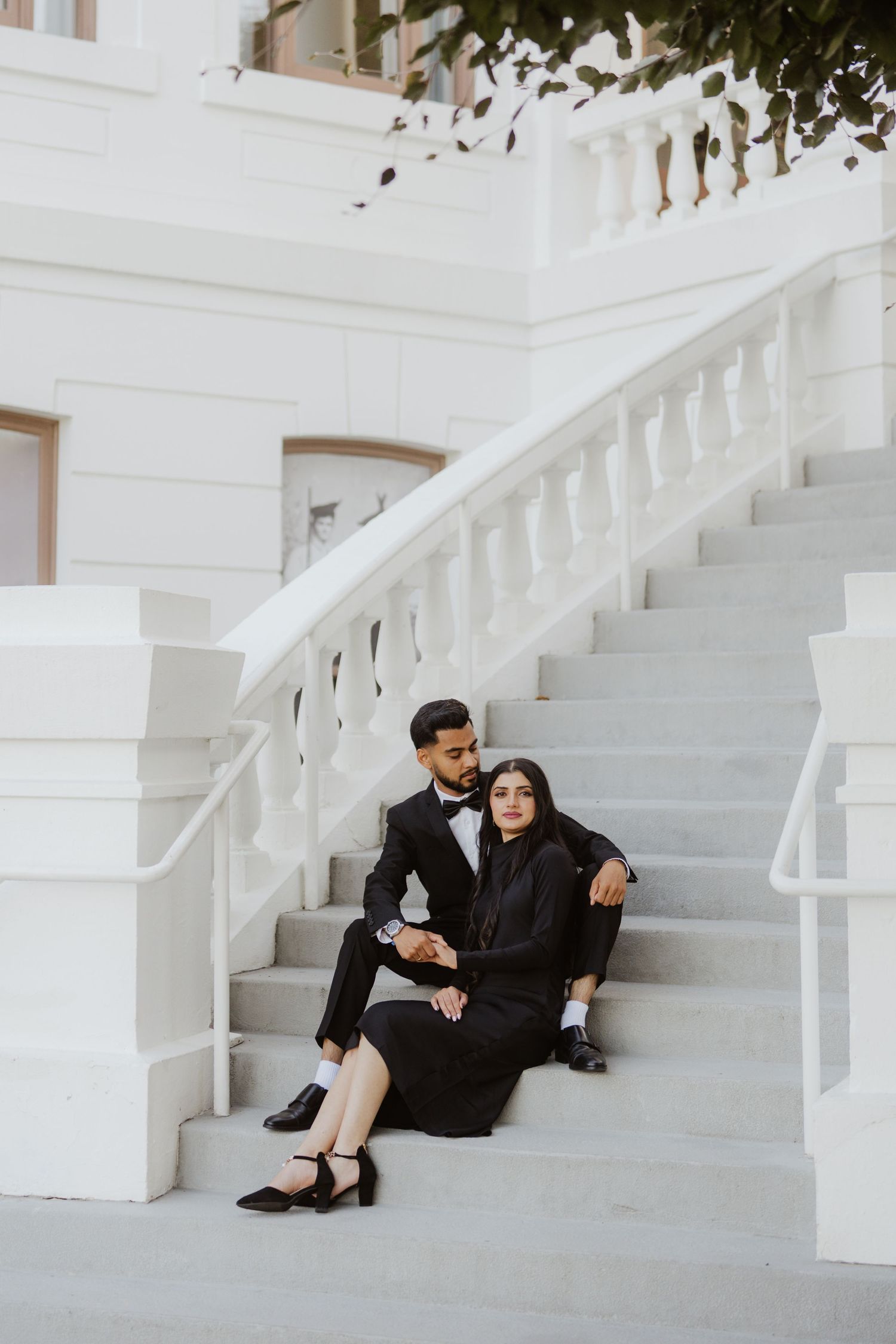 Stylish couple in black formal attire sitting together on white marble stairs of a classic building.
