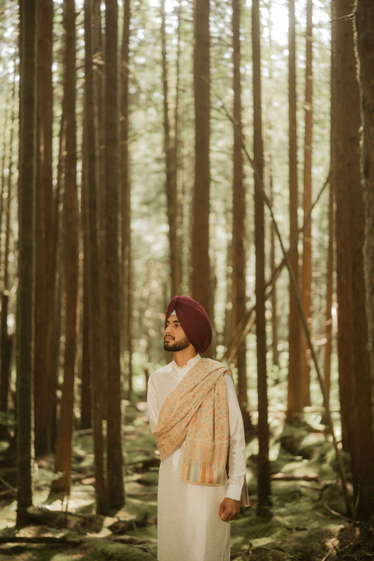 Sikh man in white kurta and red turban standing peacefully in a sunlit forest.