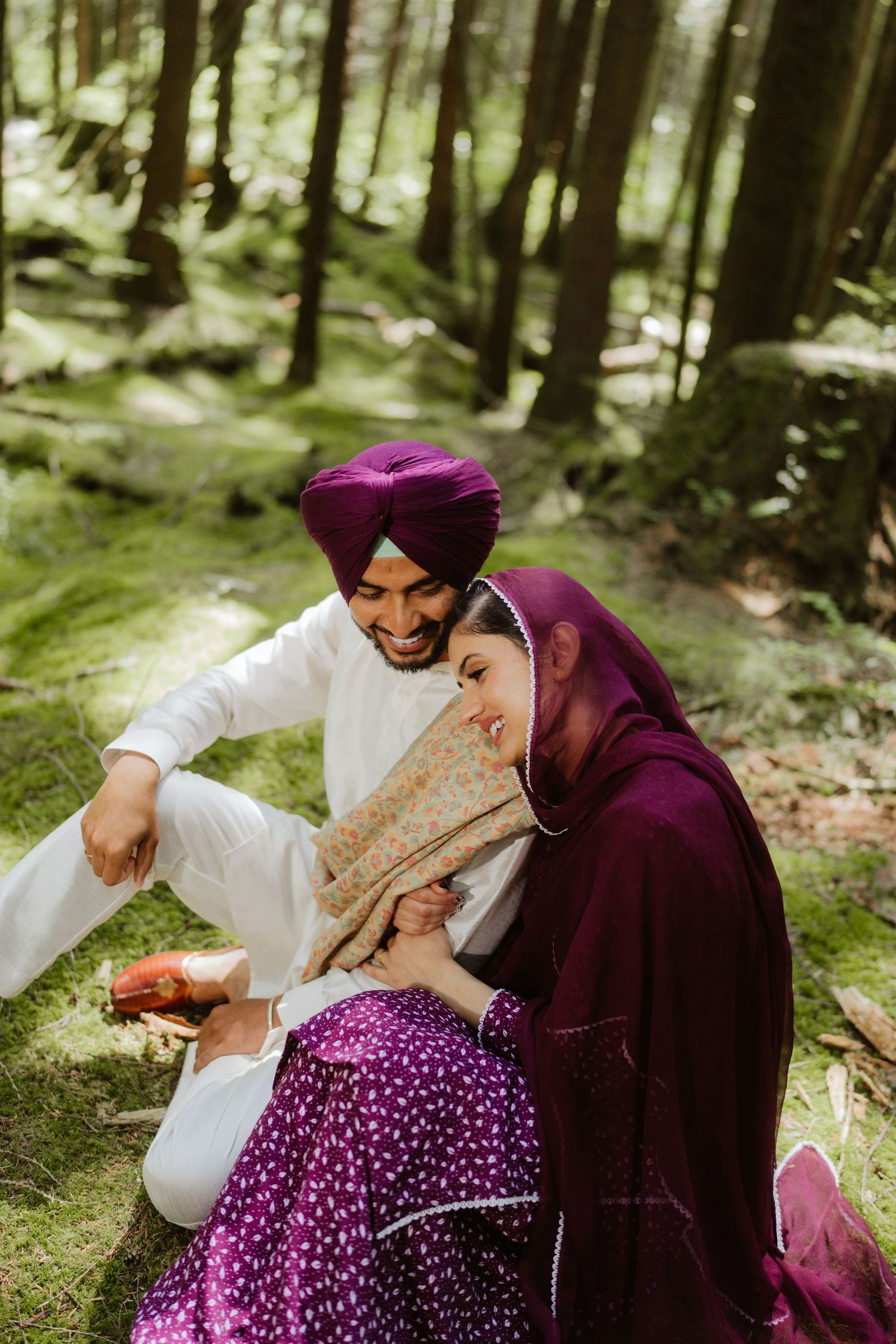Sikh couple laughing together in green forest, woman in purple dupatta leaning on man in white kurta.