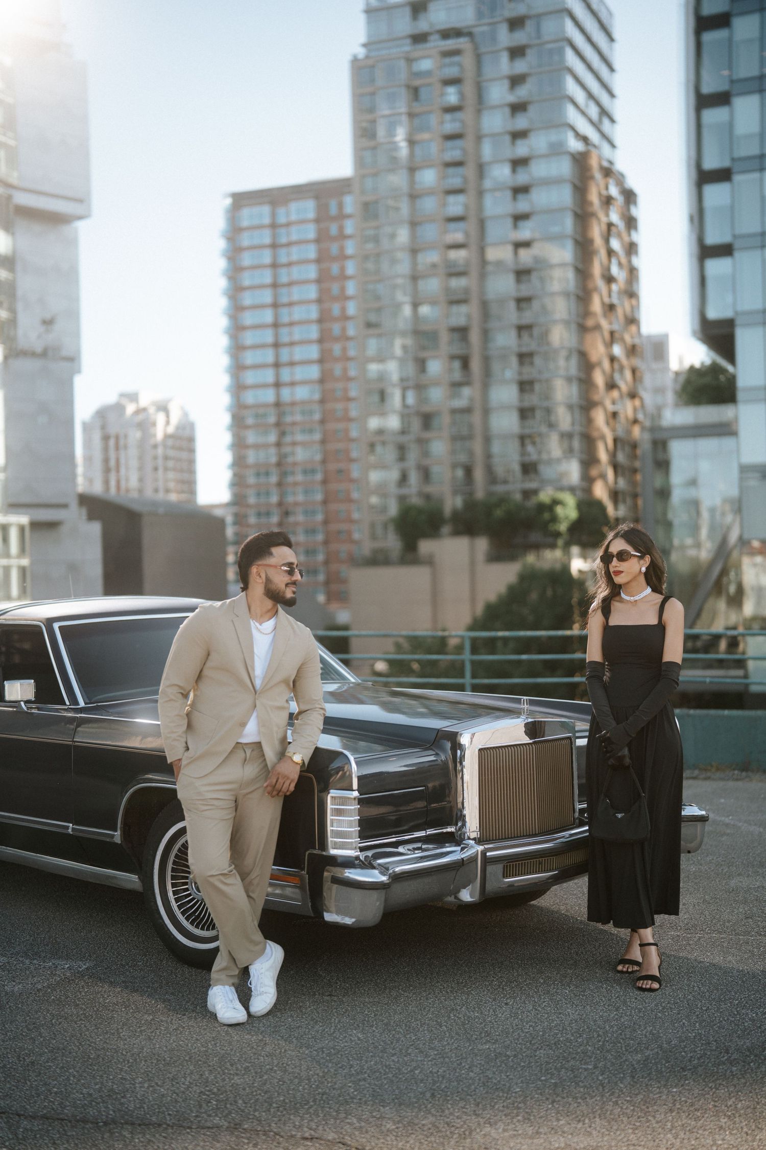 A couple poses elegantly next to a vintage black limousine in an urban setting with high-rise buildings in the background.