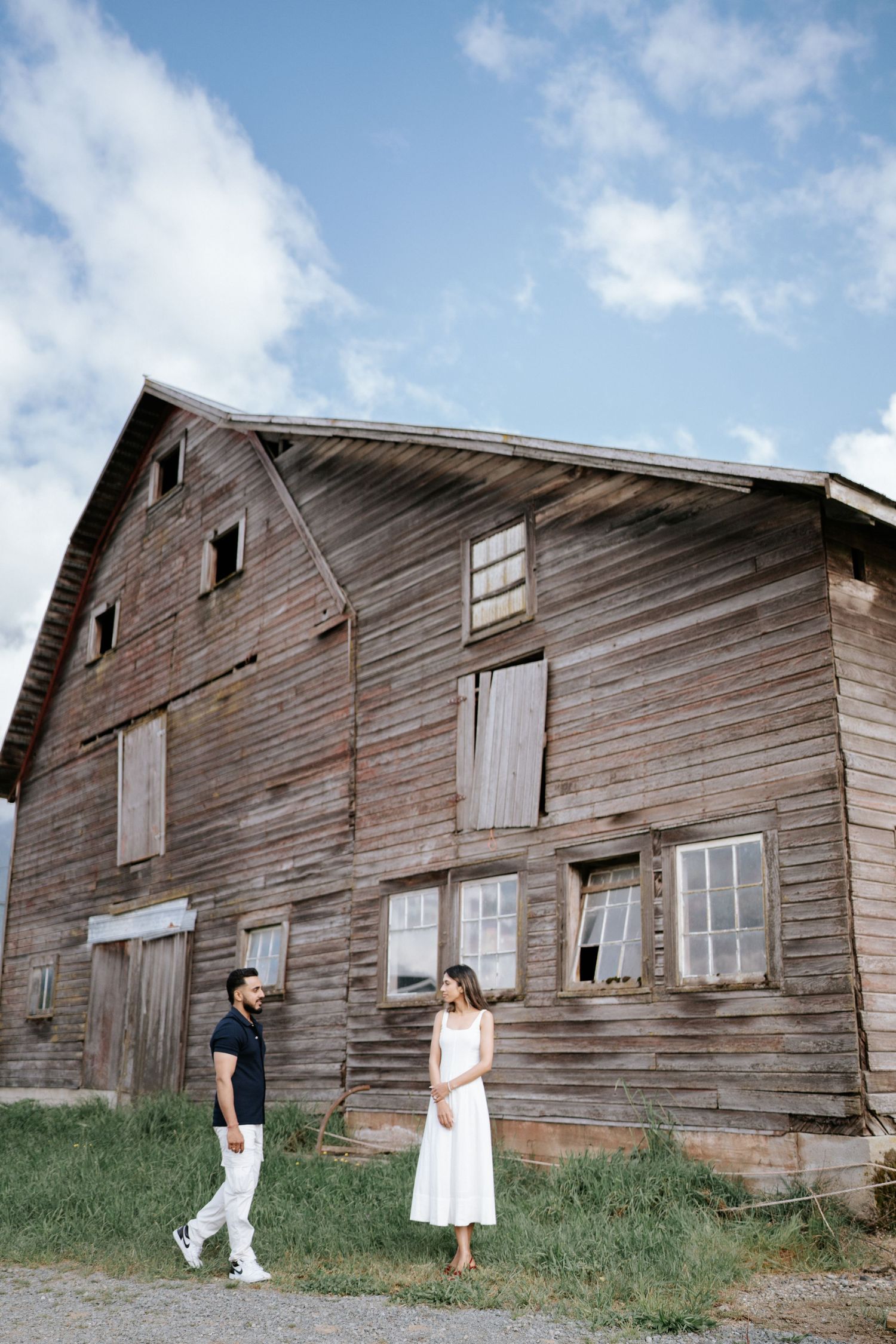 A couple stands in front of a weathered wooden barn with rustic siding and windows on a sunny day.