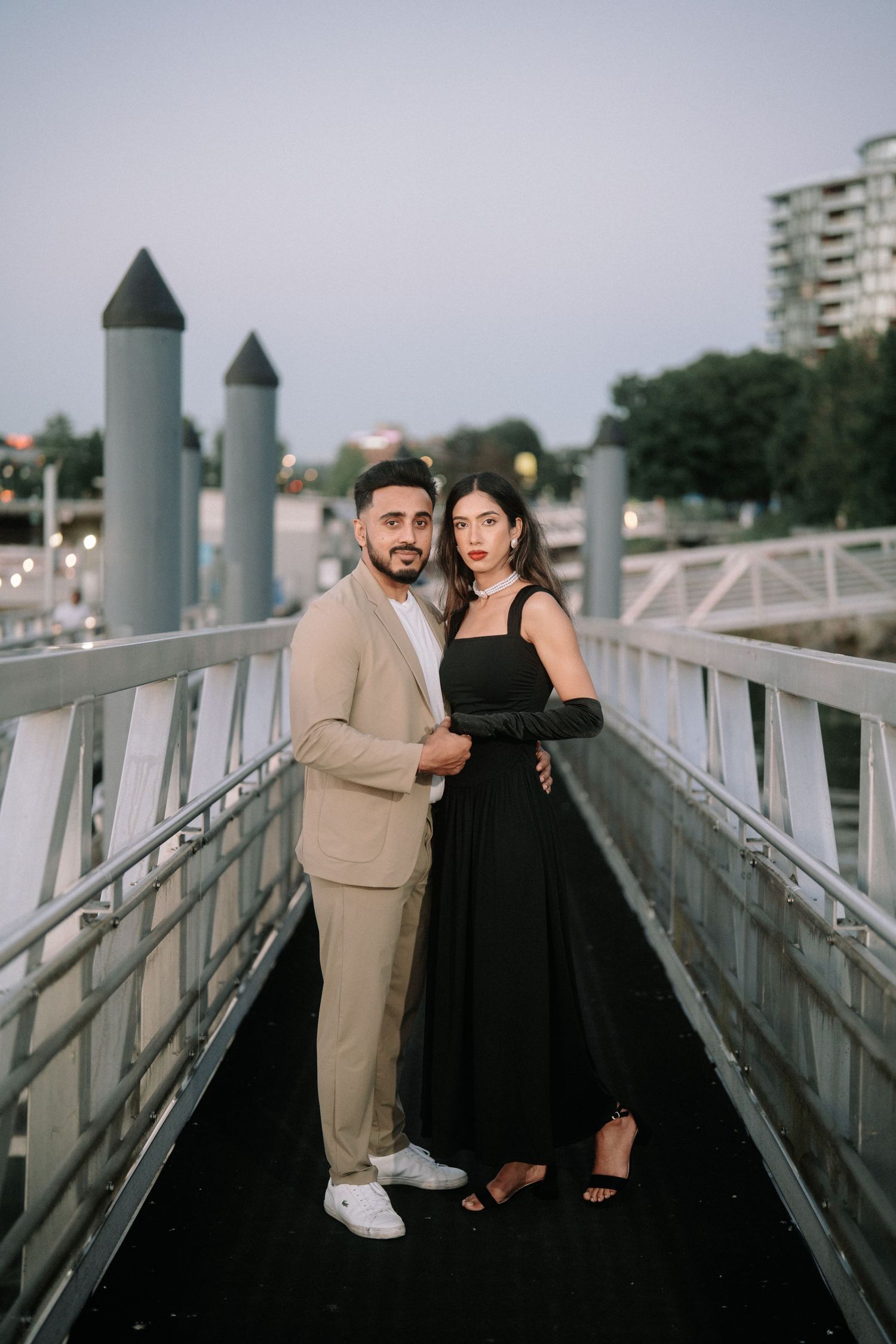 A couple poses together on a modern bridge walkway at dusk, with urban architecture visible in the background.
