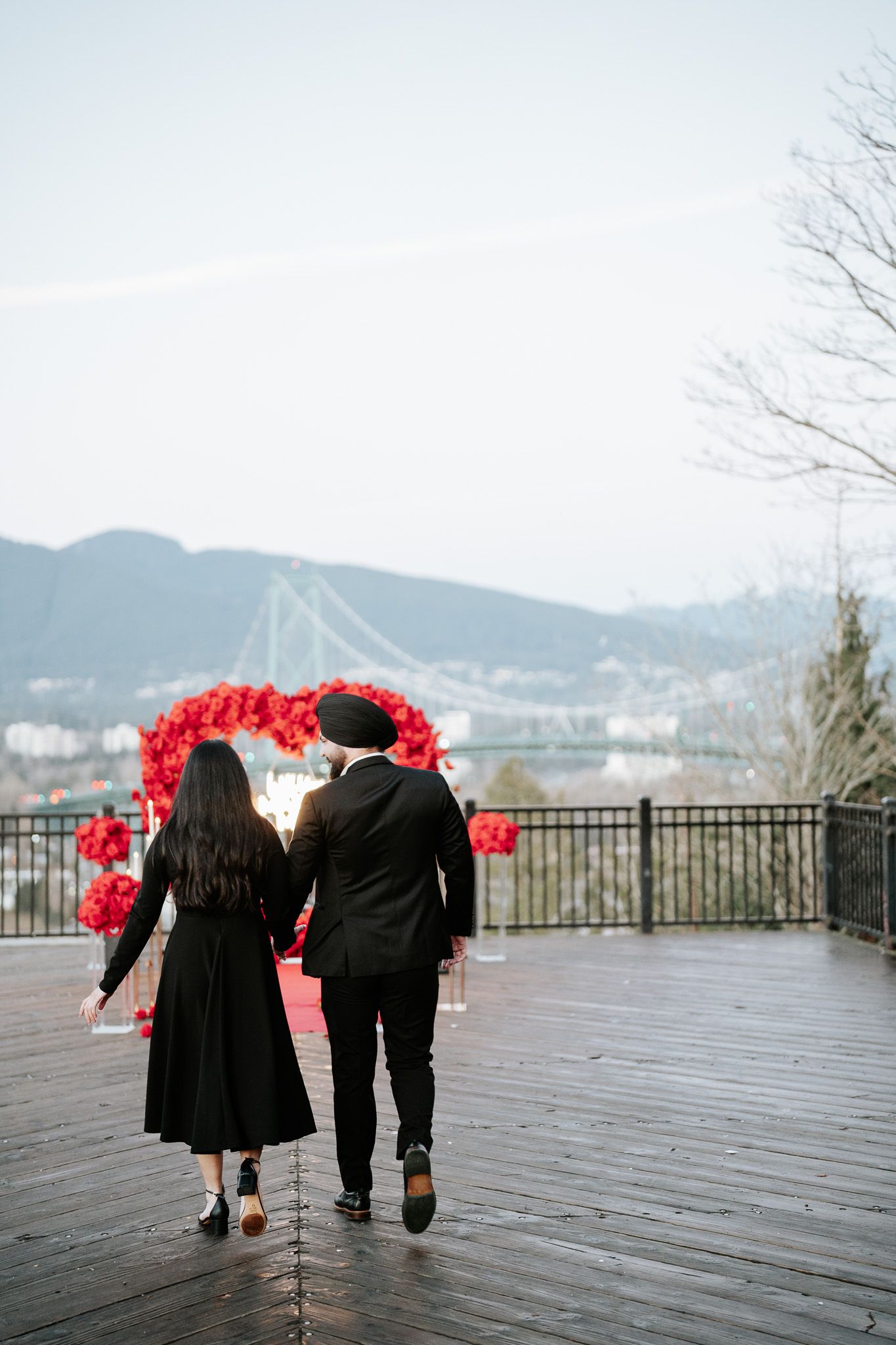 A couple in black attire walking on a wooden boardwalk with red floral decorations against a mountain backdrop.