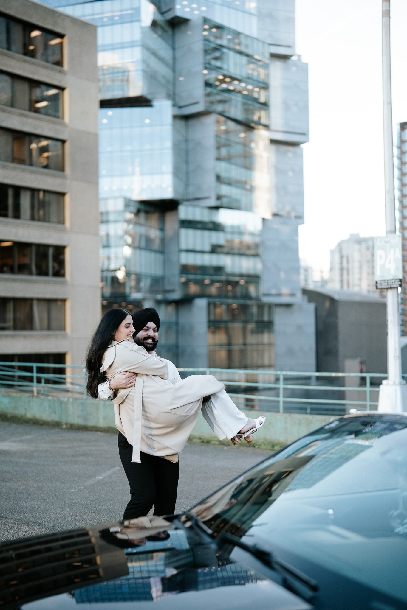 A couple embraces in a playful lift pose against an urban backdrop of modern glass buildings and parked cars.