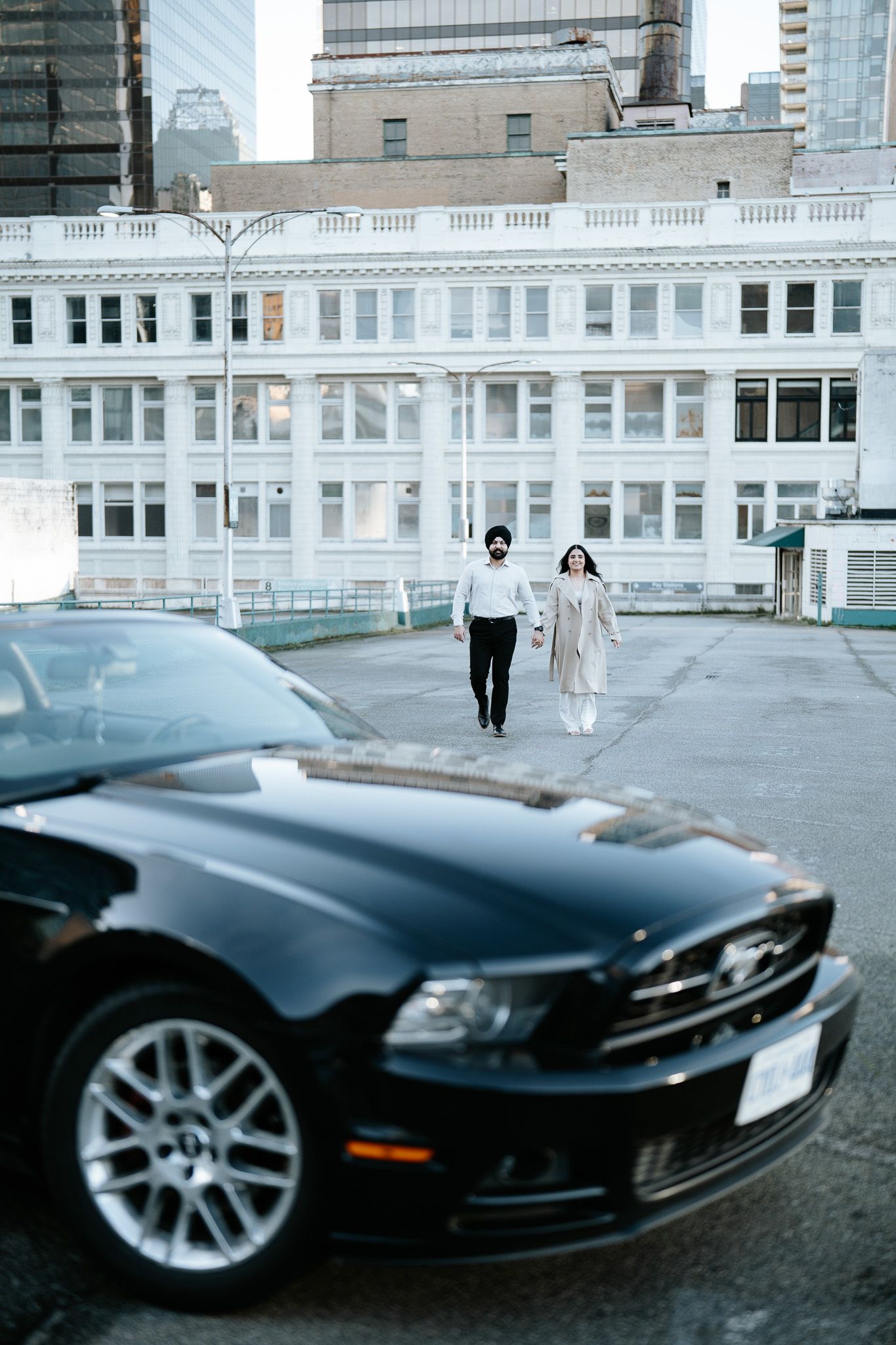 A couple walks together on a rooftop parking area with a black Ford Mustang in the foreground and city buildings behind them.