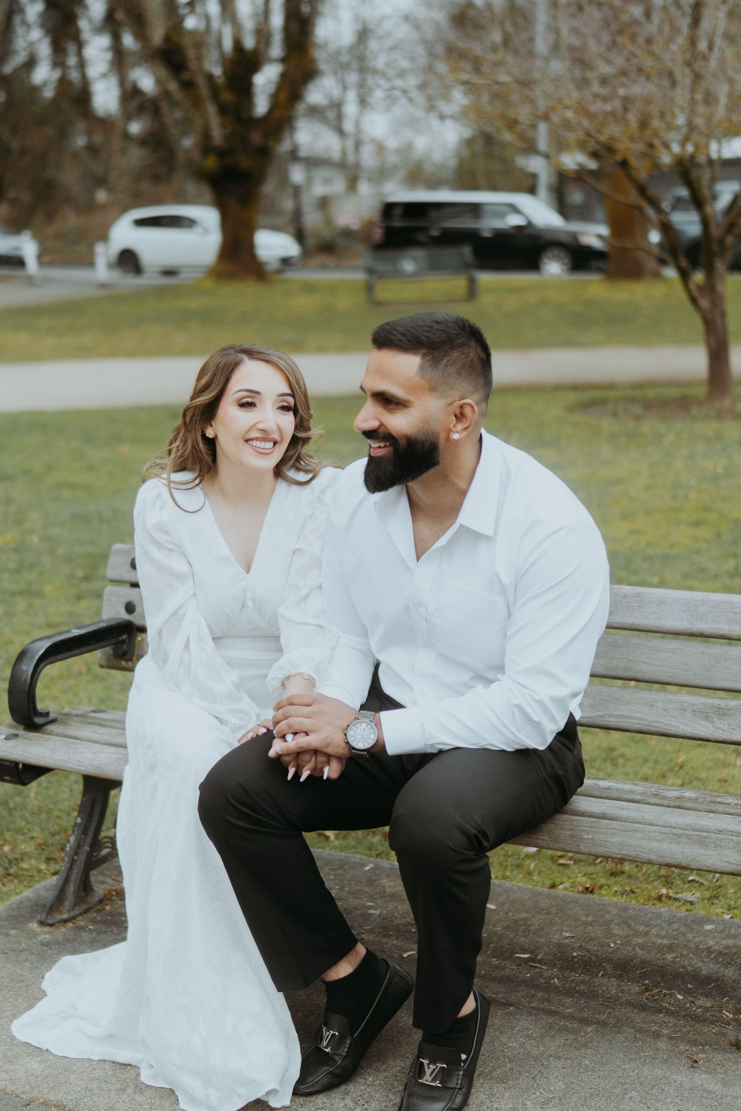 A couple in coordinated white outfits shares genuine smiles while sitting on a wooden bench.