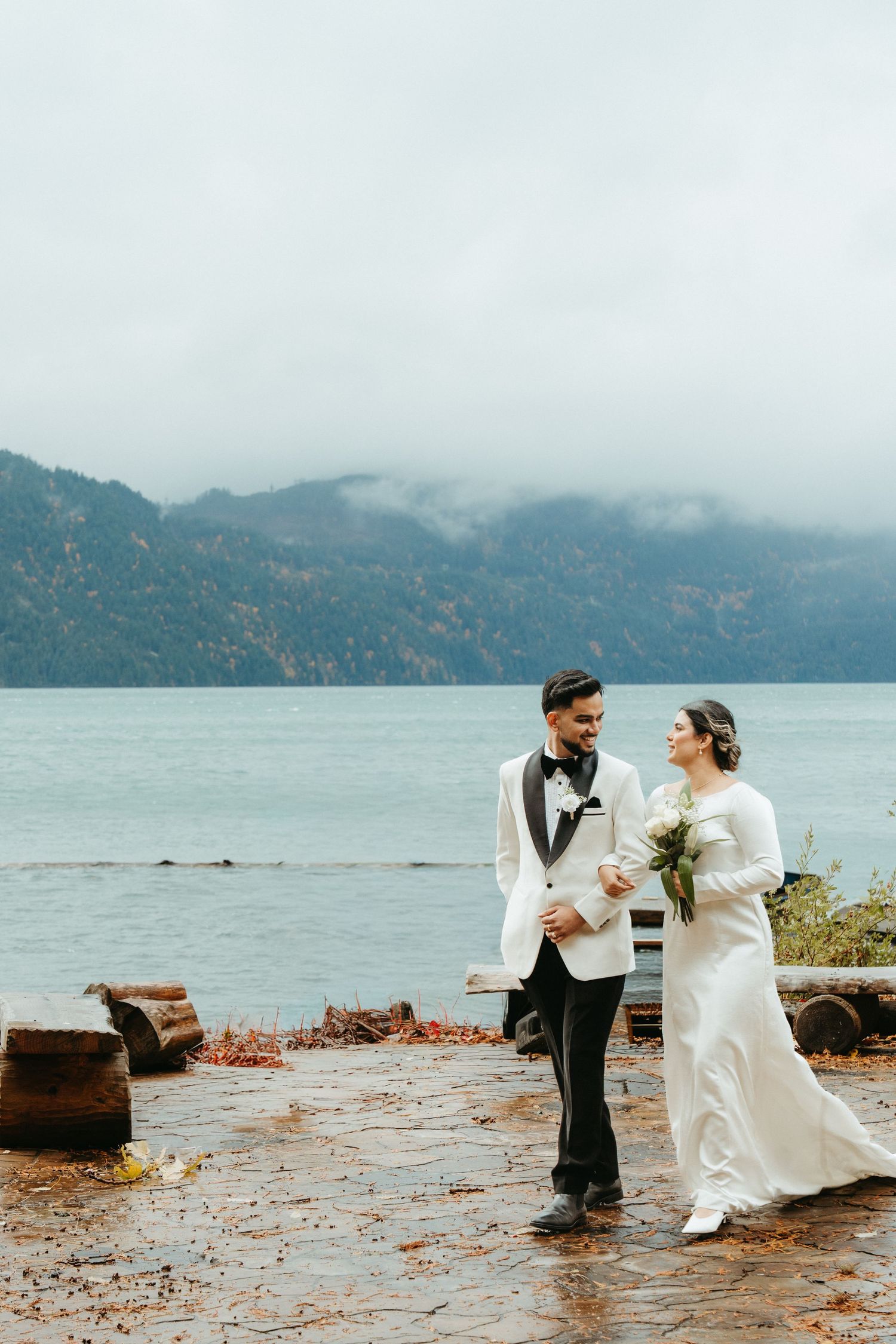 A couple shares joyful moments in their wedding attire while standing along a misty lakeside with mountains in the background.
