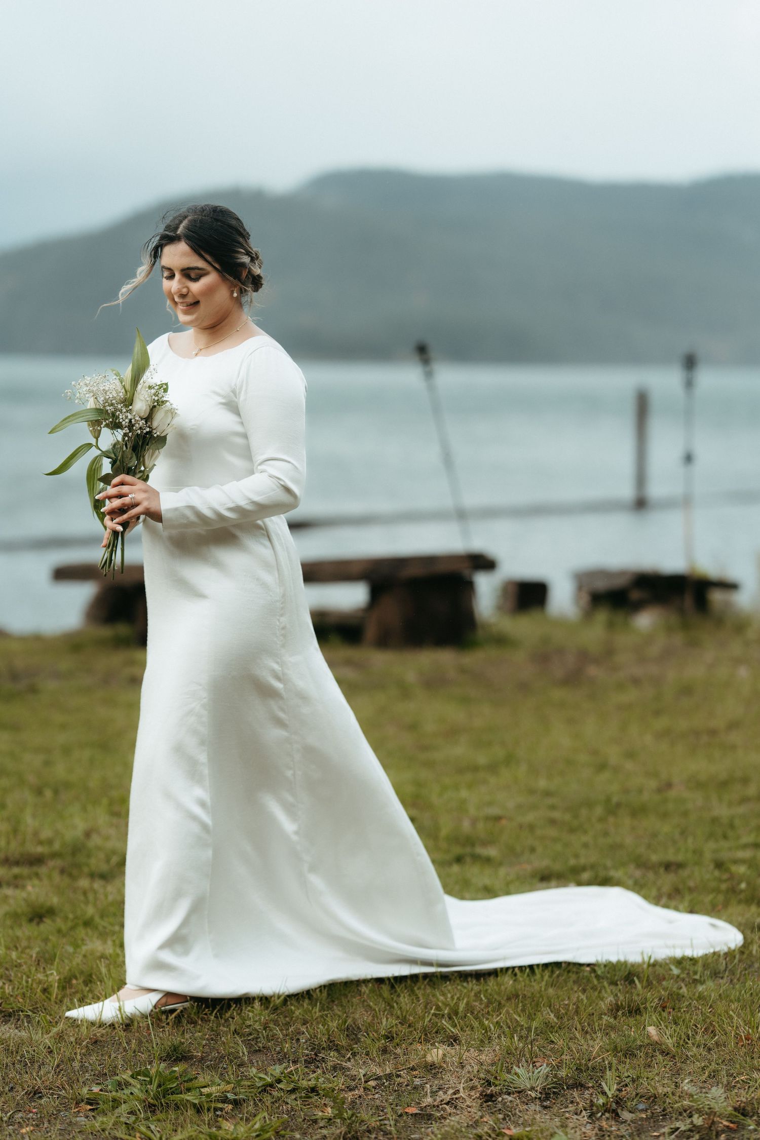 A bride in a long-sleeved white wedding dress holds a bouquet while standing on grass near a misty lake.