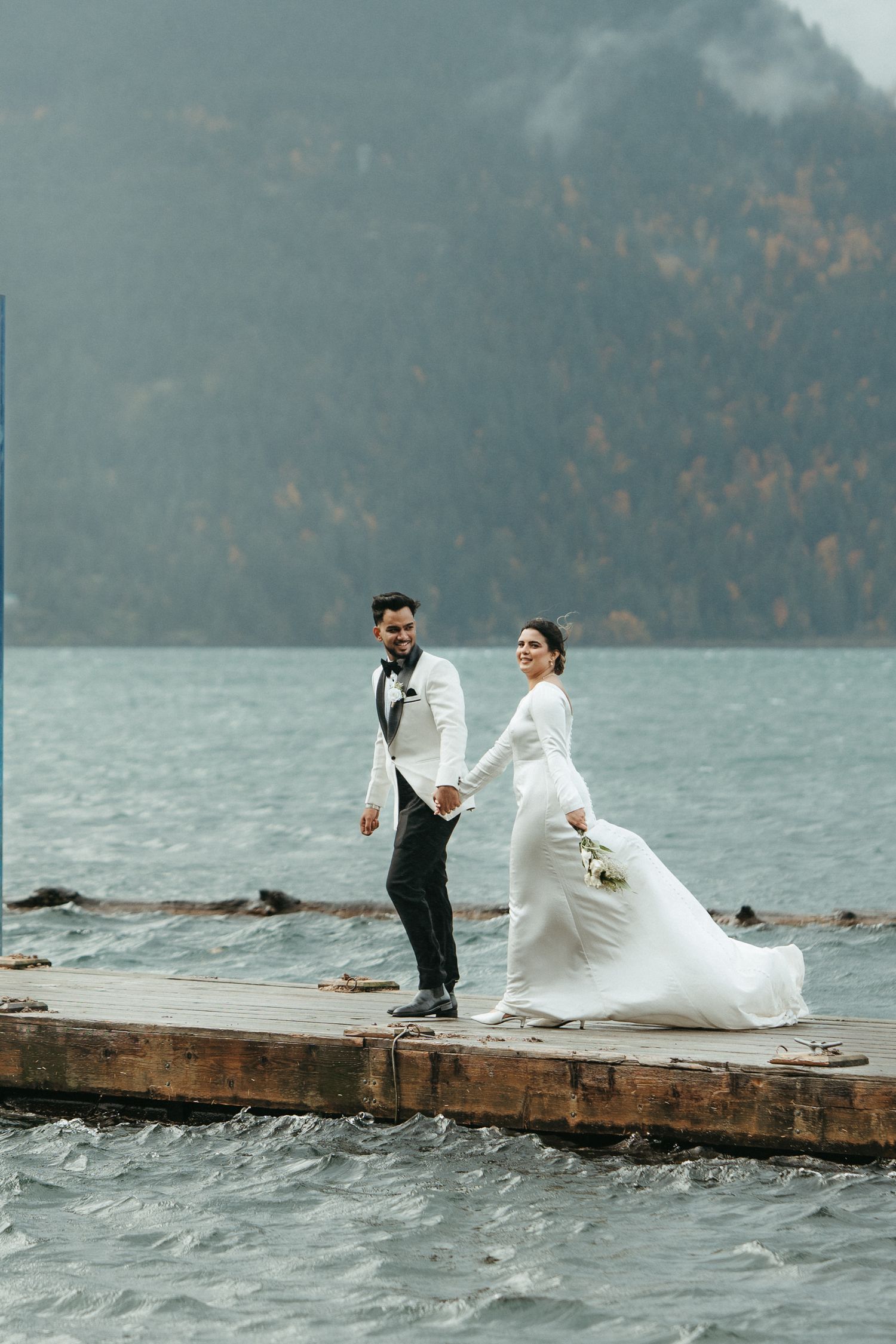 A couple in wedding attire stands on a wooden dock over stormy waters with mountains in the background.