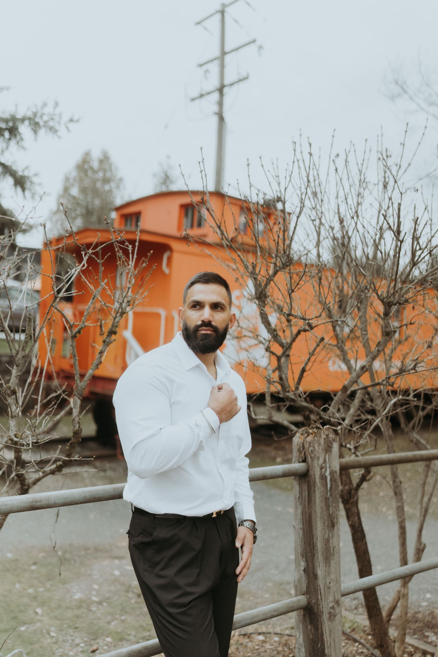 A person in a white shirt and dark pants poses against a backdrop of bare trees and an orange building.