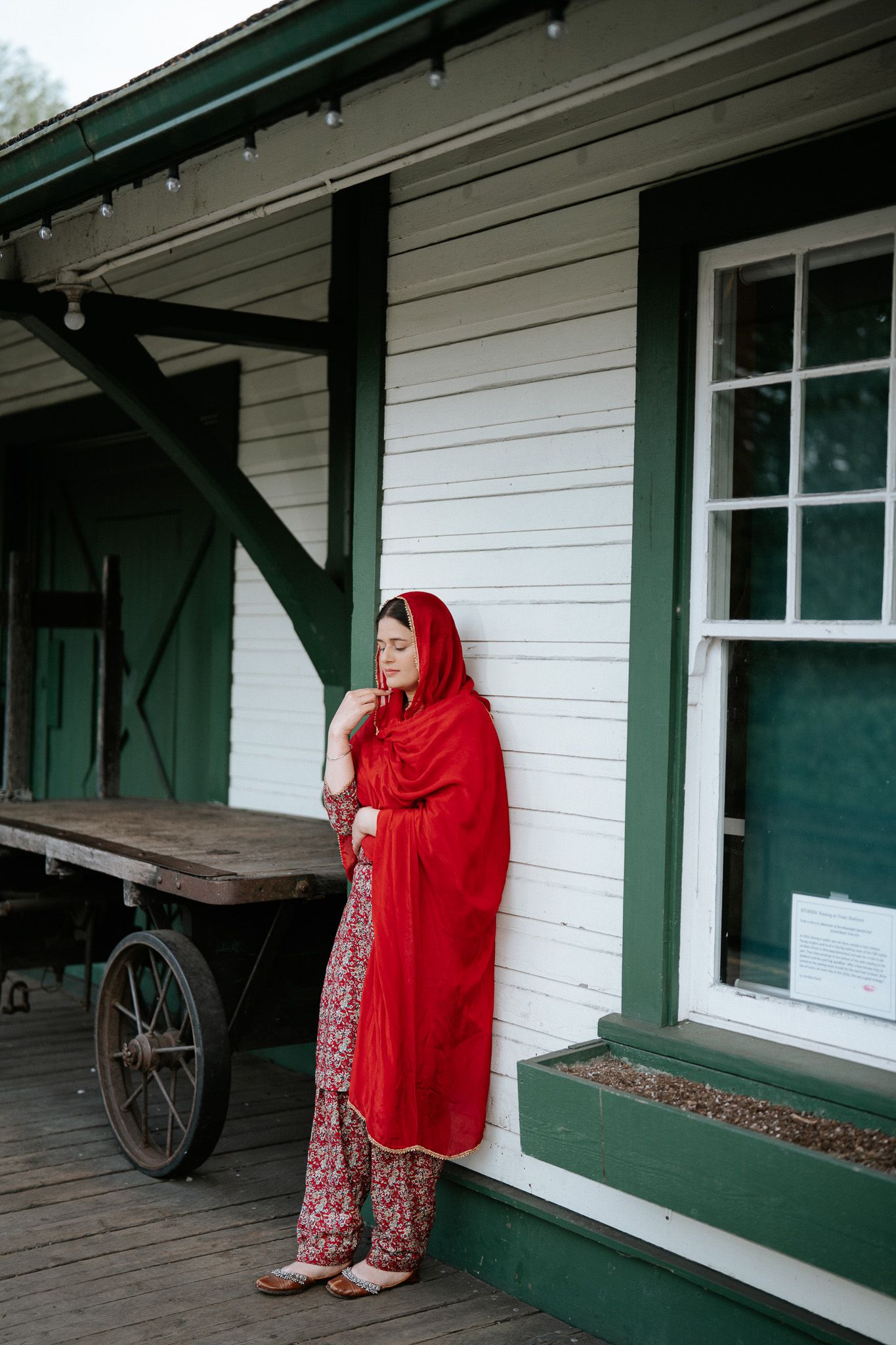 Woman in traditional red salwar with dupatta stands leaning against a white heritage building exterior.