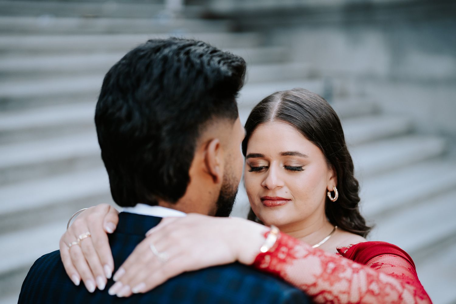 A couple in traditional South Asian attire shares an intimate embrace, the woman in red lehenga resting her head on the man's shoulder.
