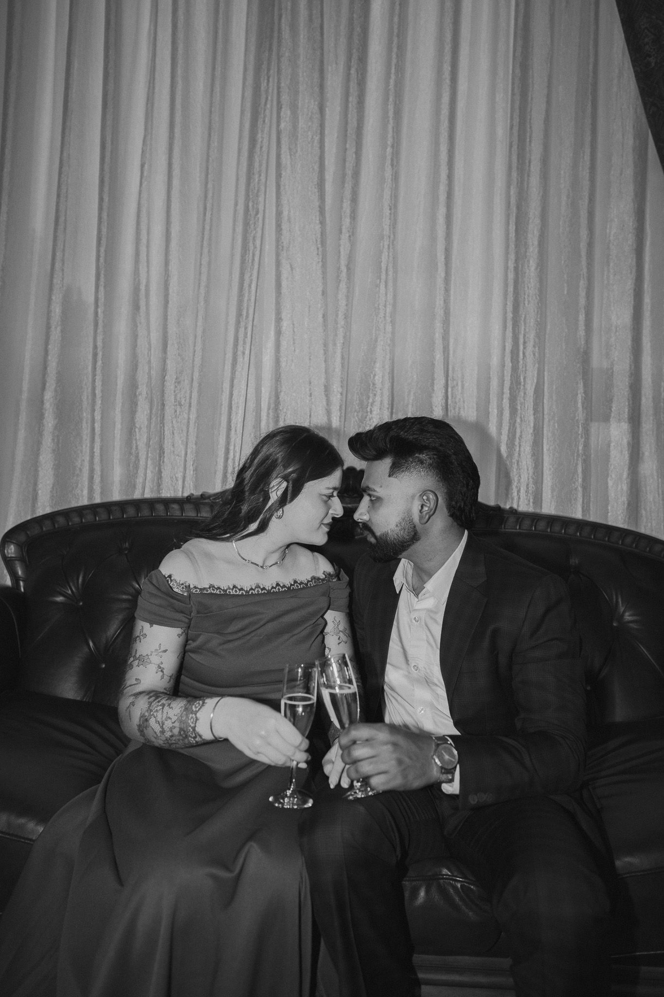 Black and white photo of romantic couple toasting with champagne on a leather sofa in formal attire.