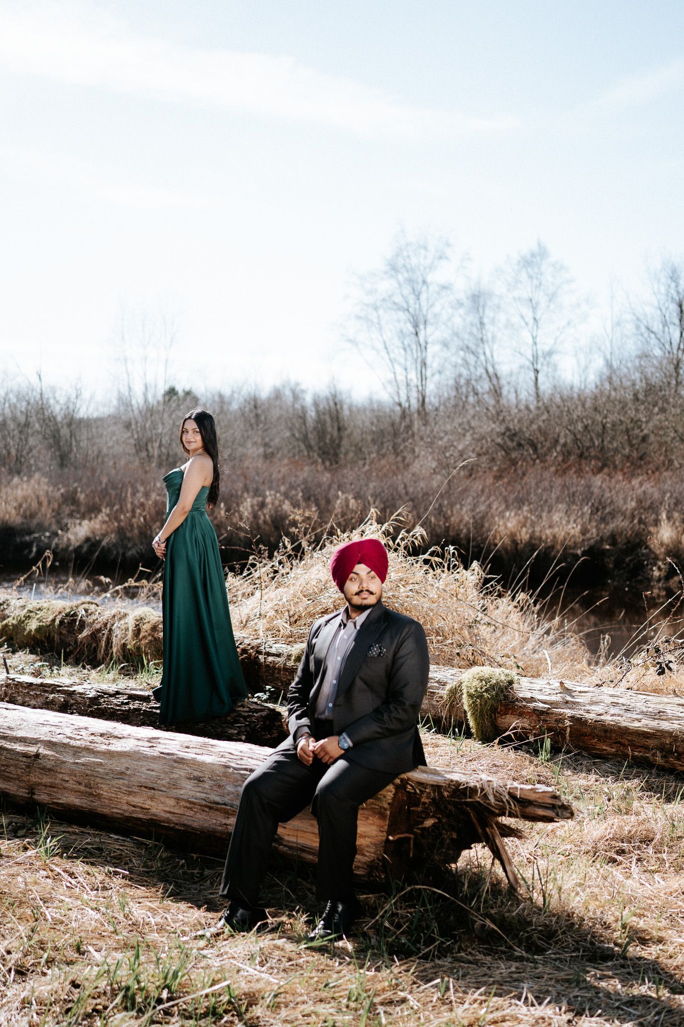 Couple sits on fallen logs in a winter field during an outdoor photoshoot session.